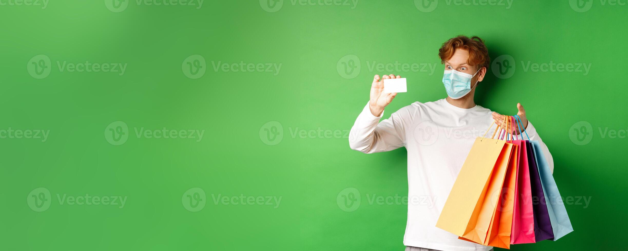 Handsome young man in medical mask, showing plastic credit card and shopping bags with items purchased with discount, green background photo