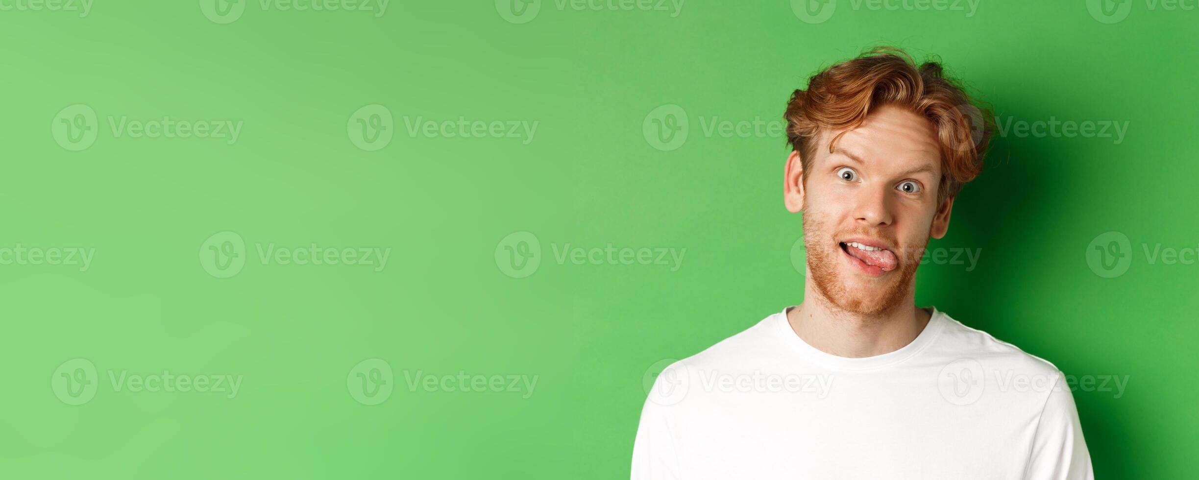 Emotions and fashion concept. Close up of funny redhead man showing silly faces, sticking tongue and staring at camera, standing over green background photo
