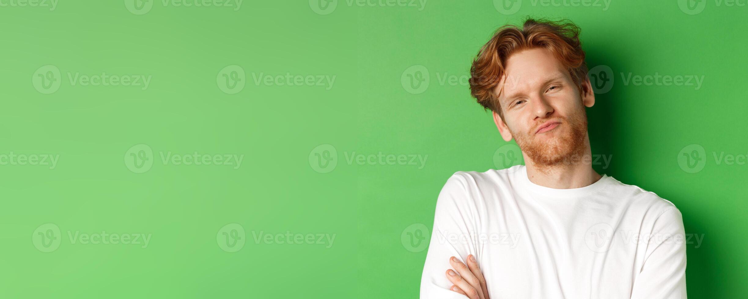 Emotions and fashion concept. Close-up of skeptical young man with red hair and beard pucker lips, looking at camera as waiting for something, standing over green background photo