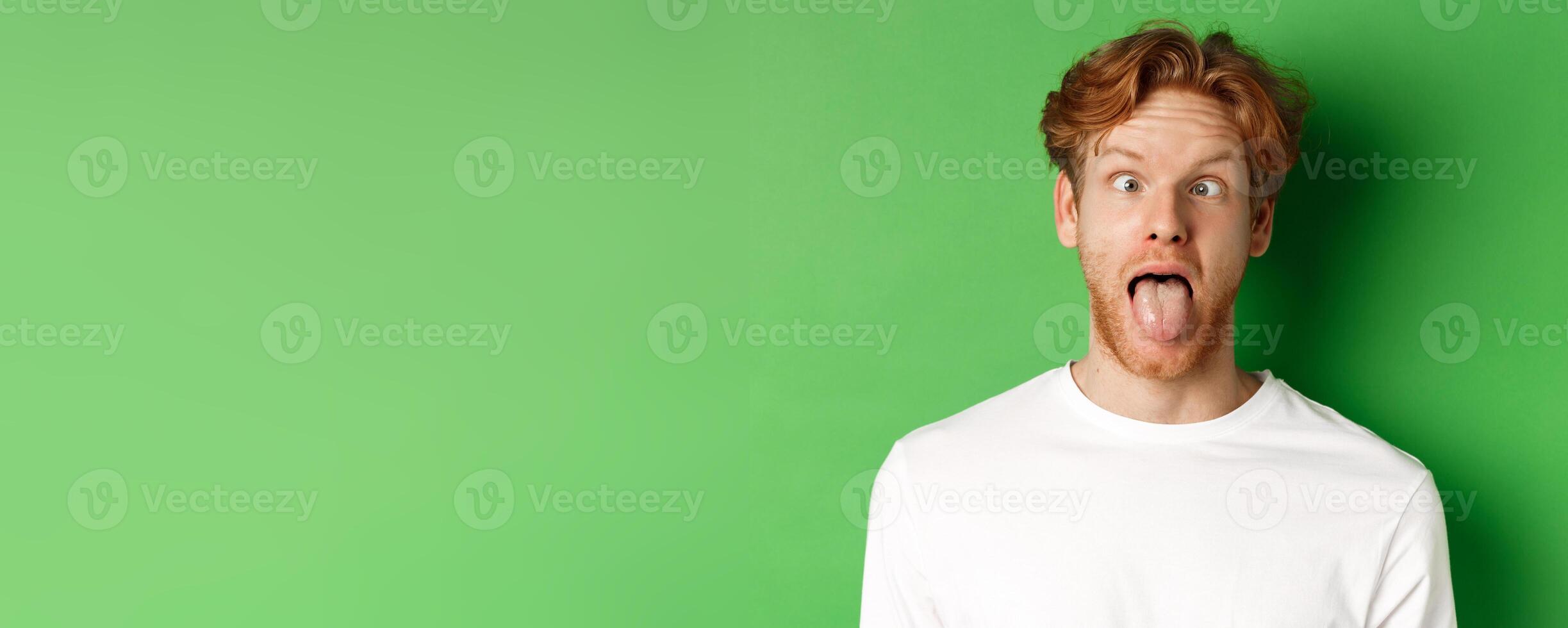 Emotions and fashion concept. Close up of funny redhead man showing silly faces, sticking tongue and squinting, standing over green background photo