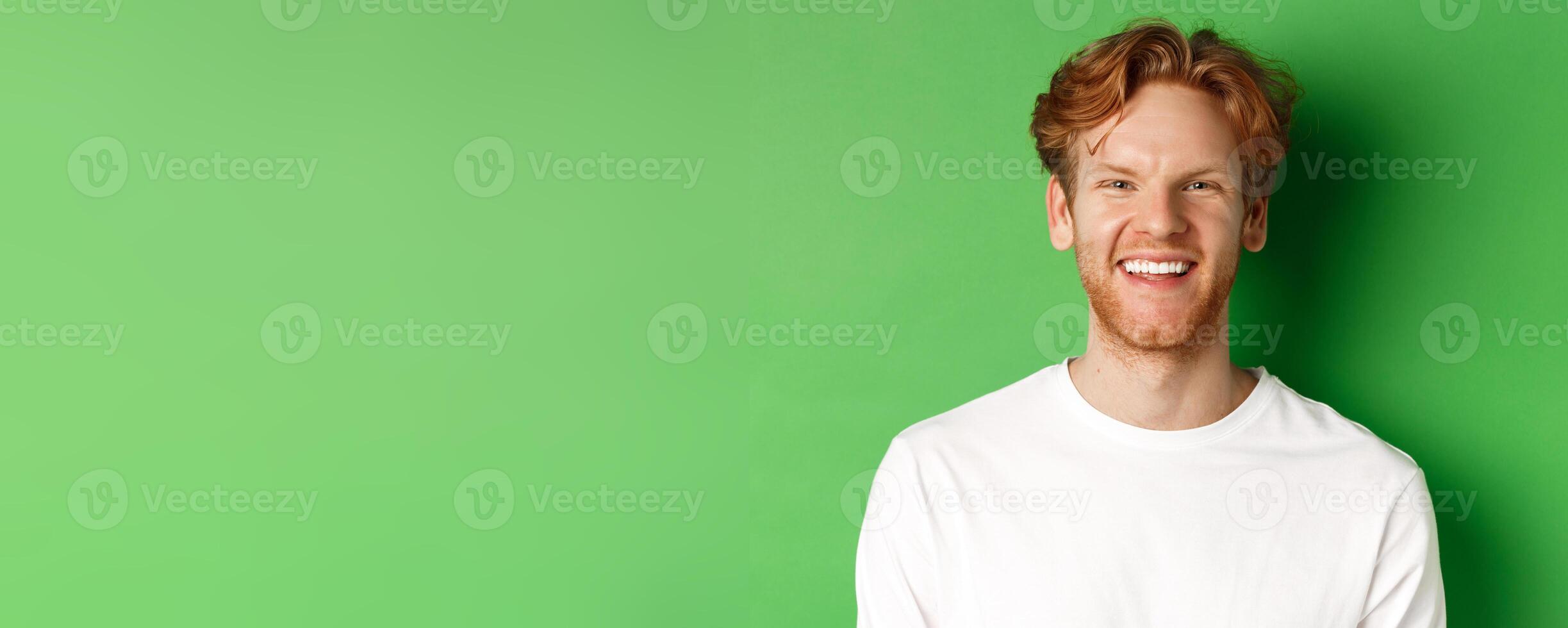 Emotions and fashion concept. Happy young man with red hair and beard, smiling and laughing at camera, standing over green background photo