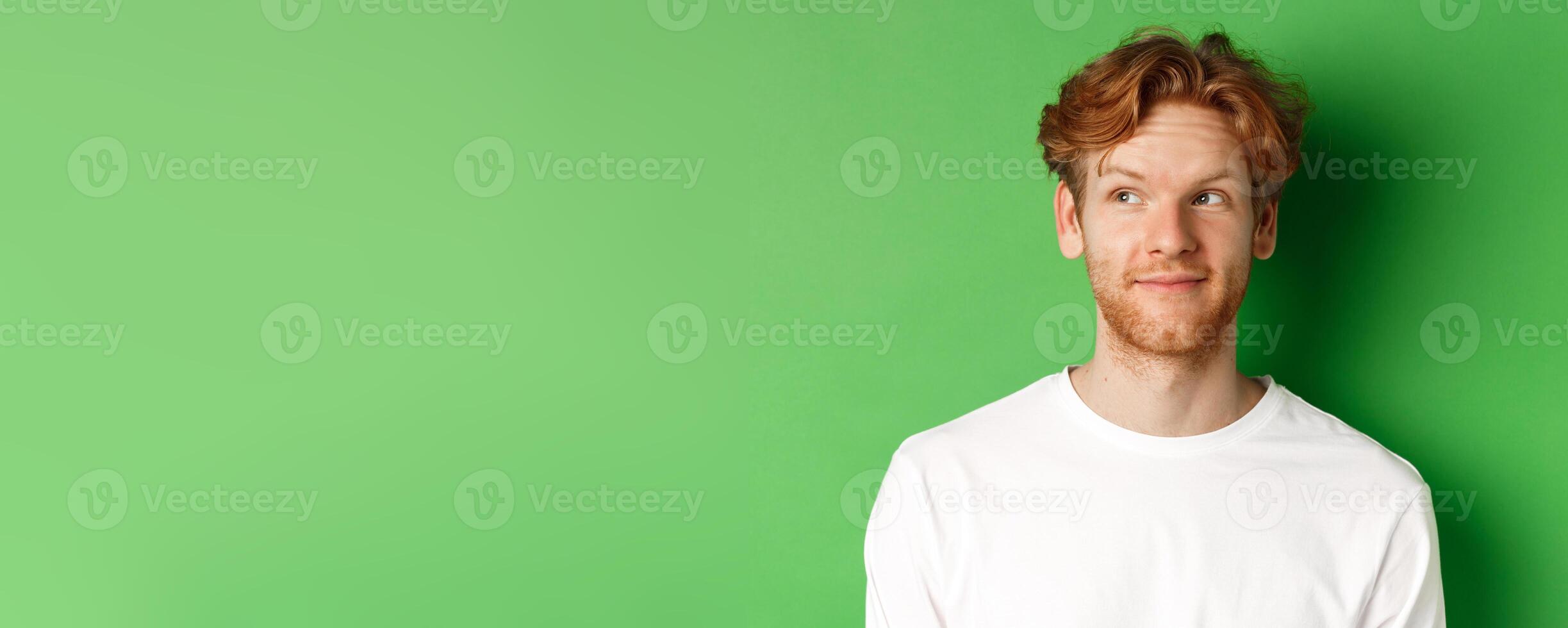 Emotions and fashion concept. Close-up of handsome redhead male model with beard and messy haircut, looking left and smiling, green background photo
