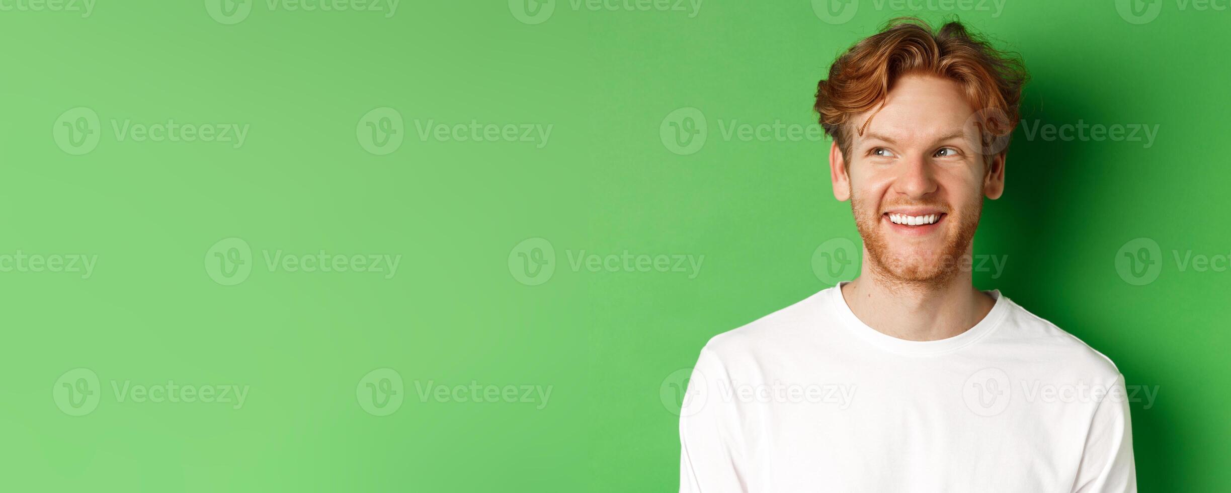 Headshot of happy redhead man with beard wearing white long sleeve, looking left at copy space and smiling, standing over green background photo