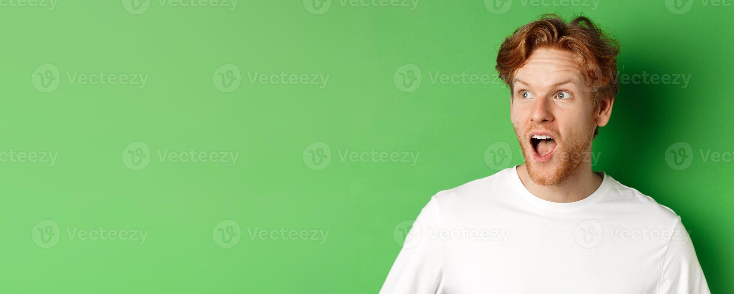Close up of surprised and impressed redhead man checking out promotion offer, looking left with dropped jaw, standing over green background photo