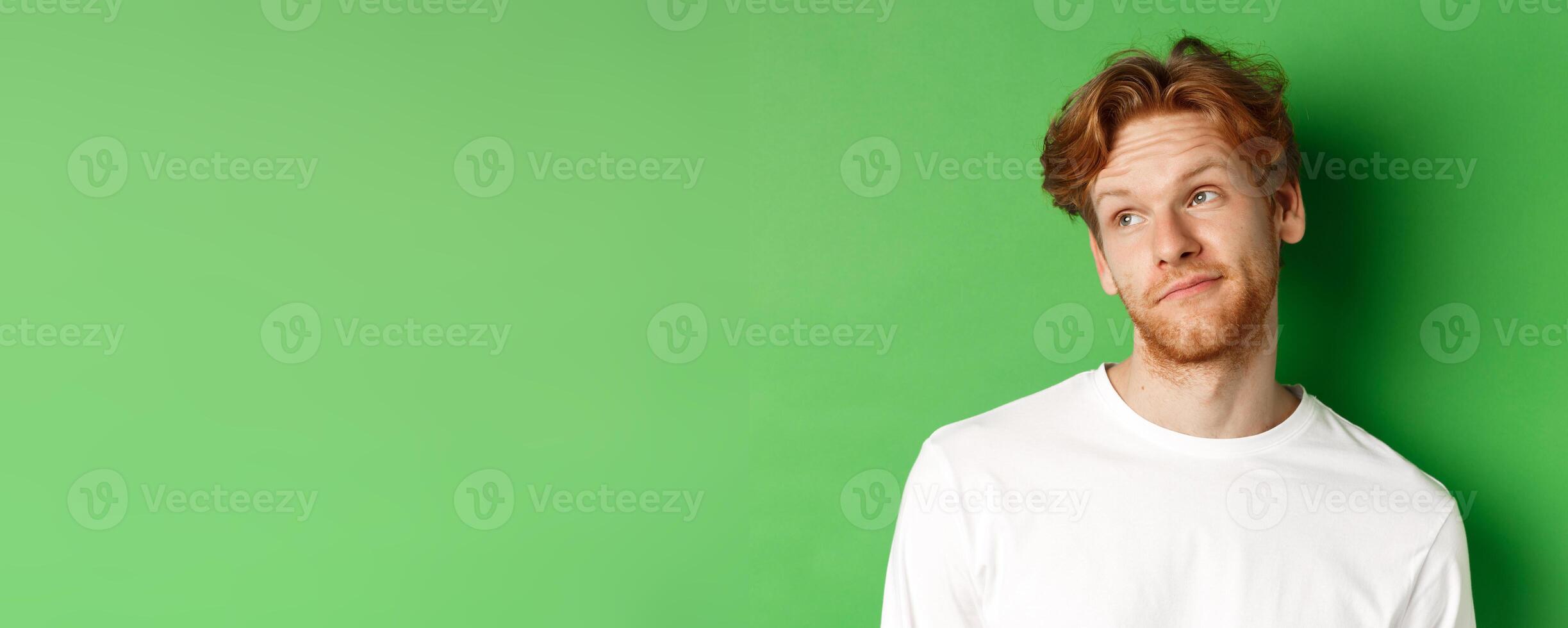 Smiling redhead man with messy haircut and beard tilt head, looking left with pleased face, standing over green background photo