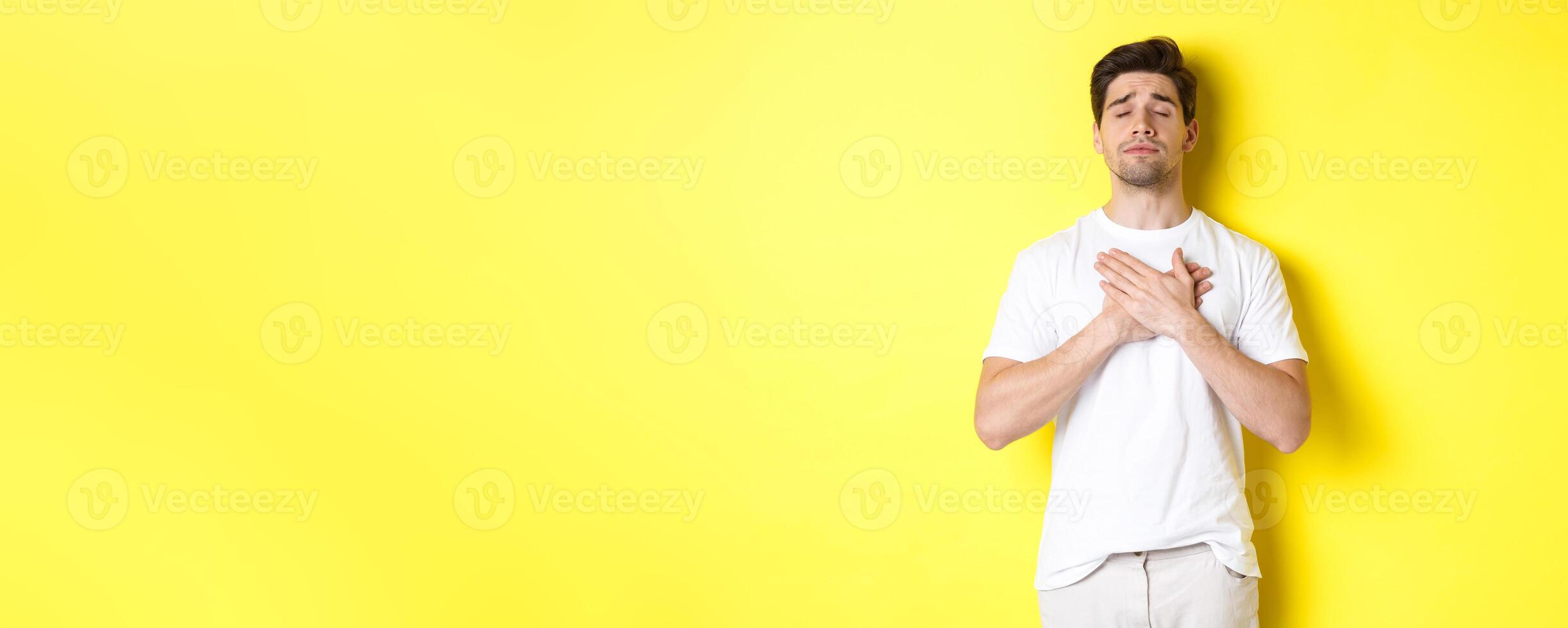 Romantic and nostalgic man holding hands on heart, close eyes and remember something, standing over yellow background photo