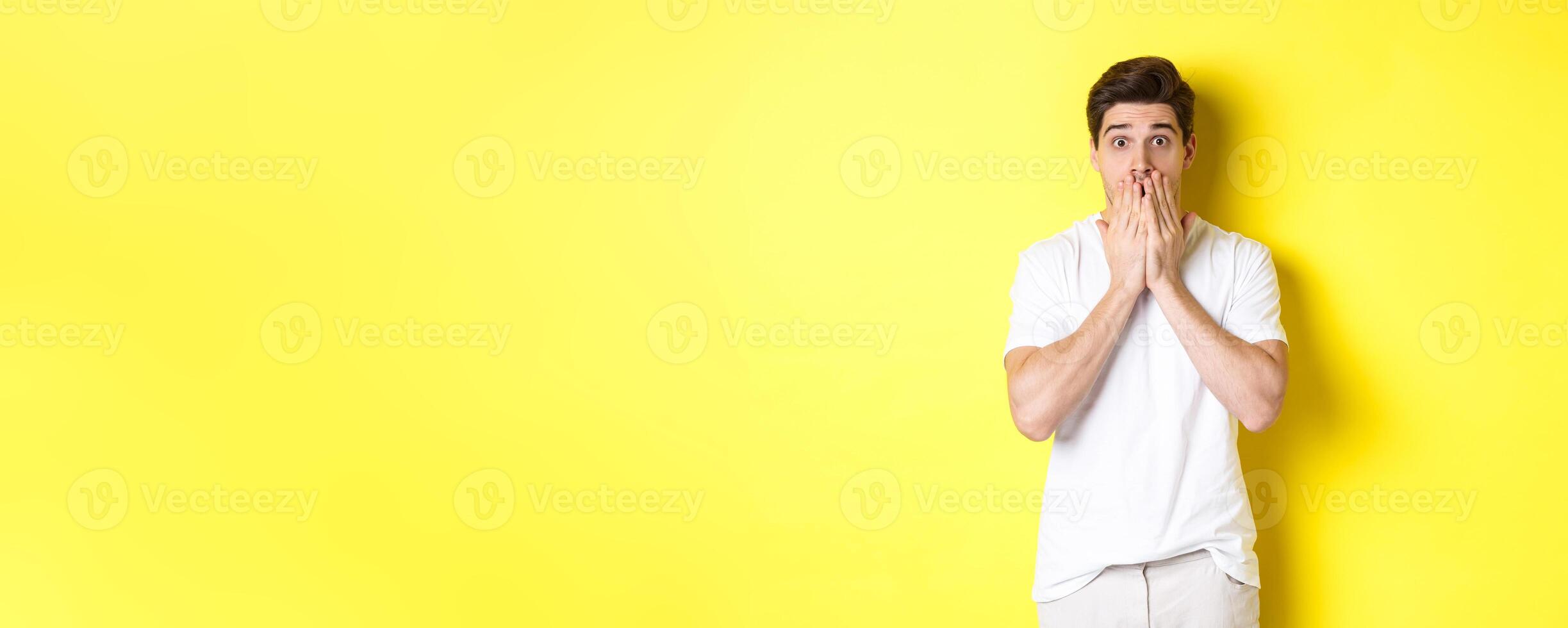 Handsome man looking shocked and speechless, holding hands on mouth, standing over yellow background photo