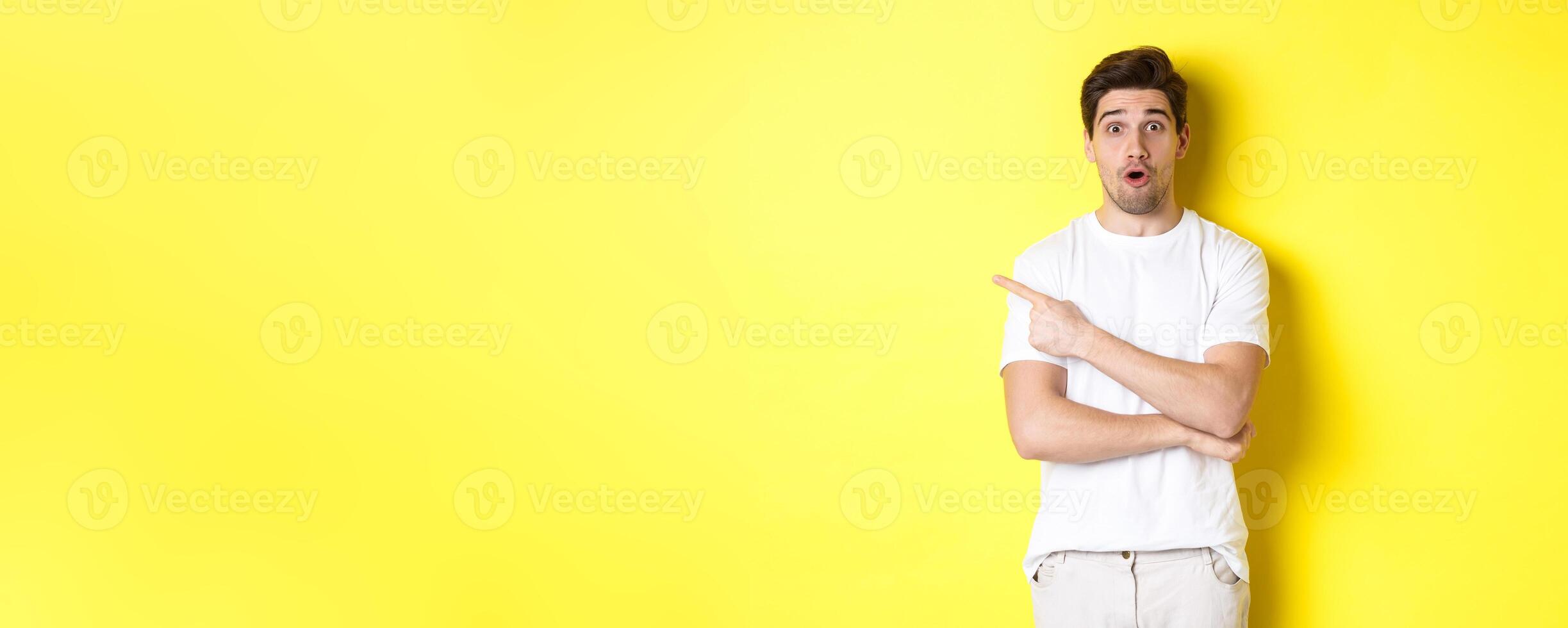 Cuious guy pointing finger left, asking question about promo offer, showing banner, standing amazed against yellow background photo