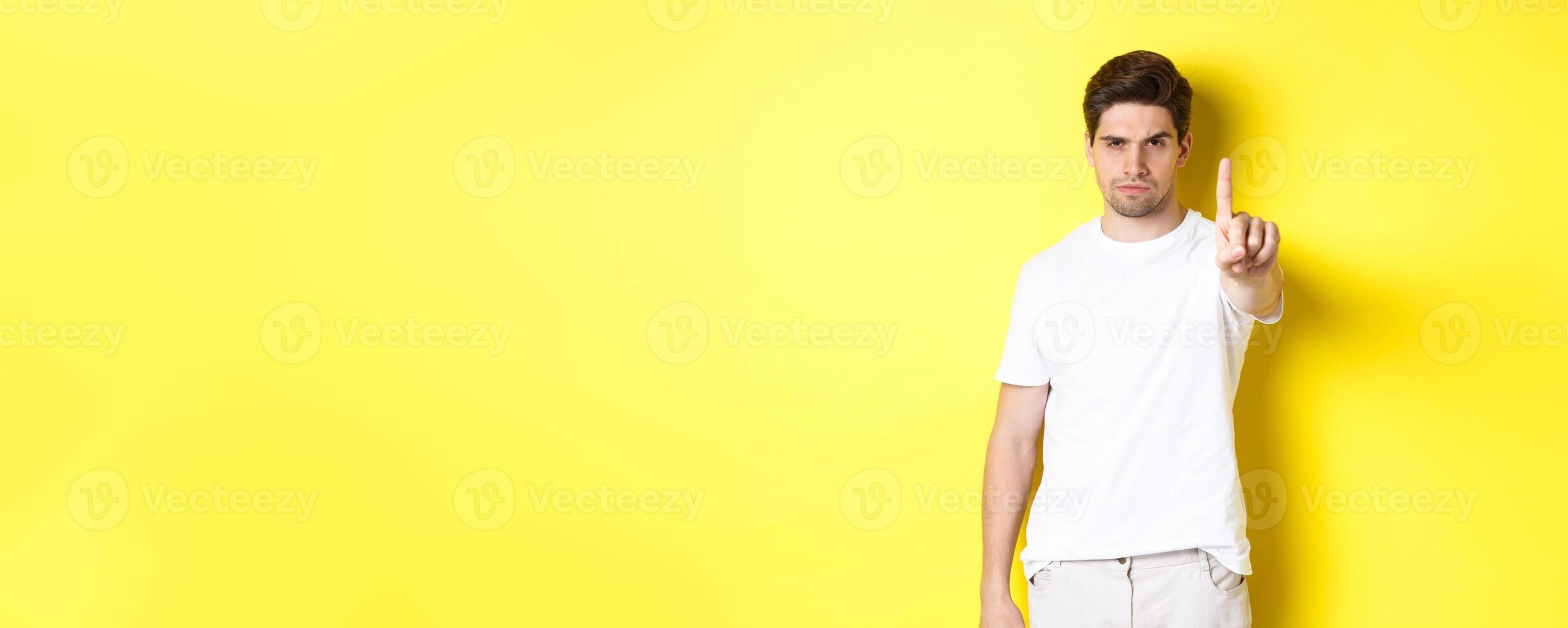 Serious man frowning displeased, shaking one finger to prohibit something, warn you, standing over yellow background photo