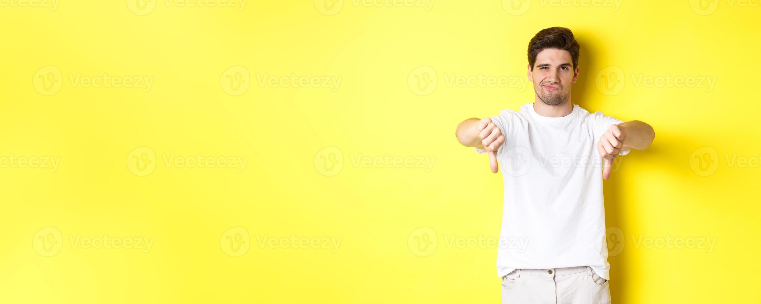 Skeptical guy showing thumbs down and smirking, dislike and disagree, standing over yellow background photo