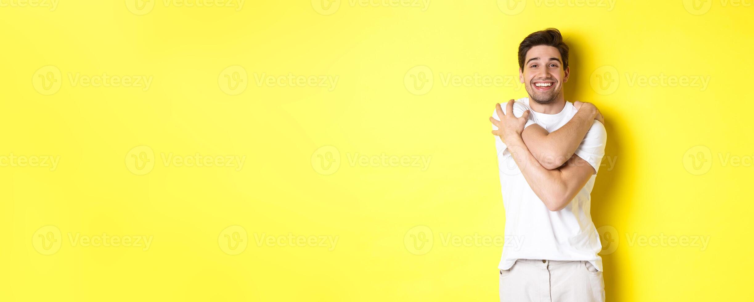 Happy kind guy hugging himself and smiling, standing pleased against yellow background photo