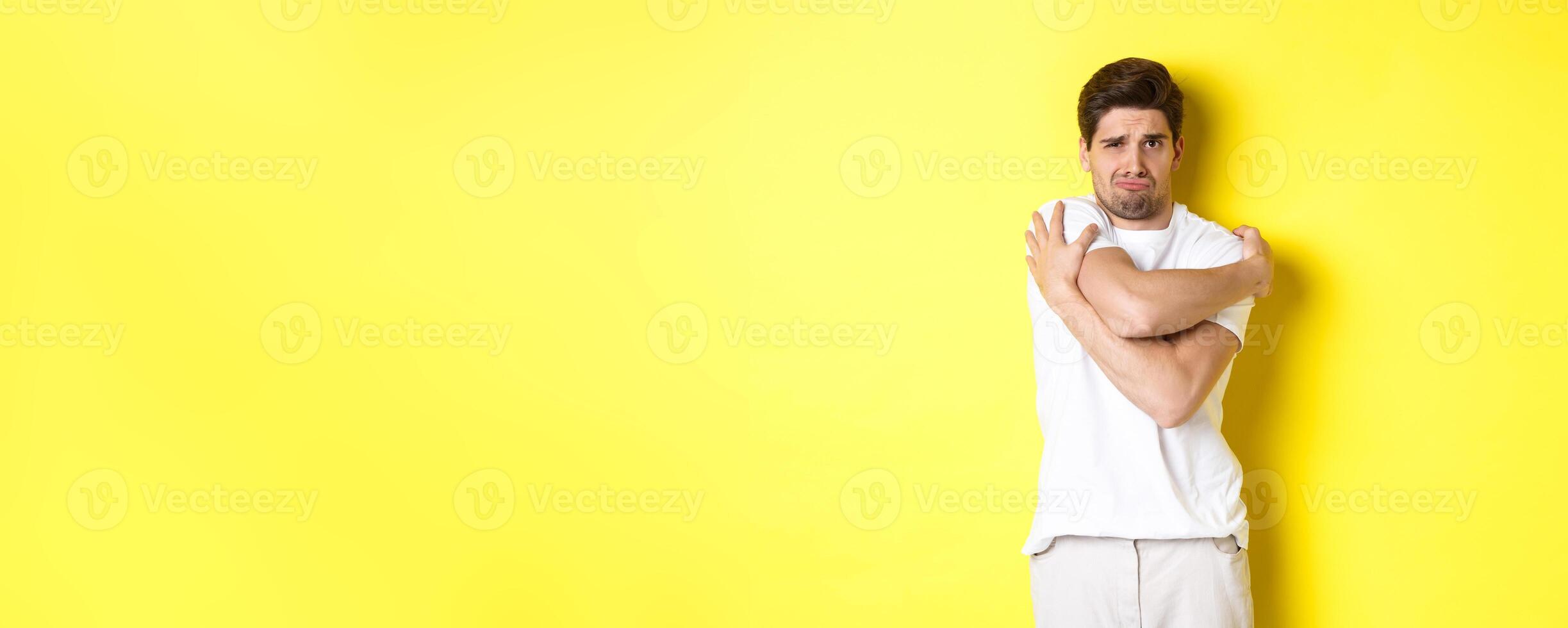Silly and timid guy trying to comfort himself, hugging his body and frowning scared, standing over yellow background photo