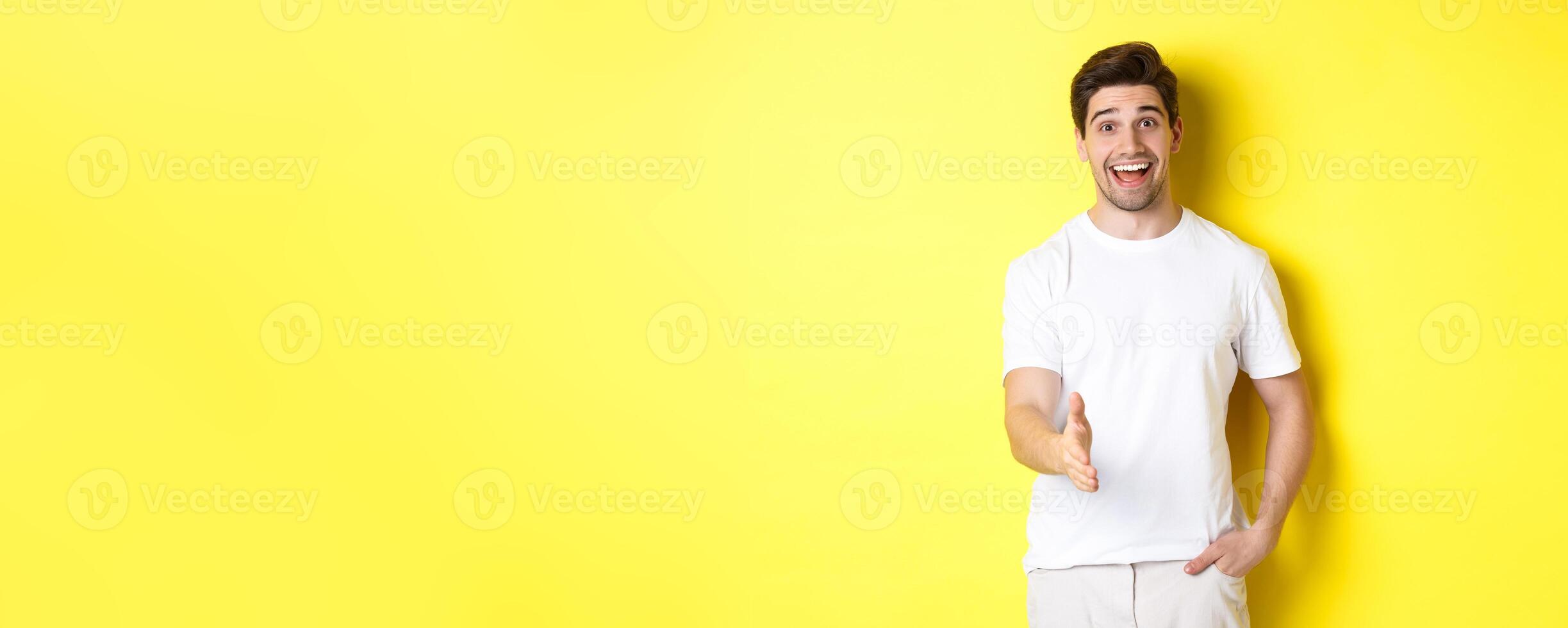 Friendly man greeting you with handshake, smiling amused, saying hello, standing over yellow background photo