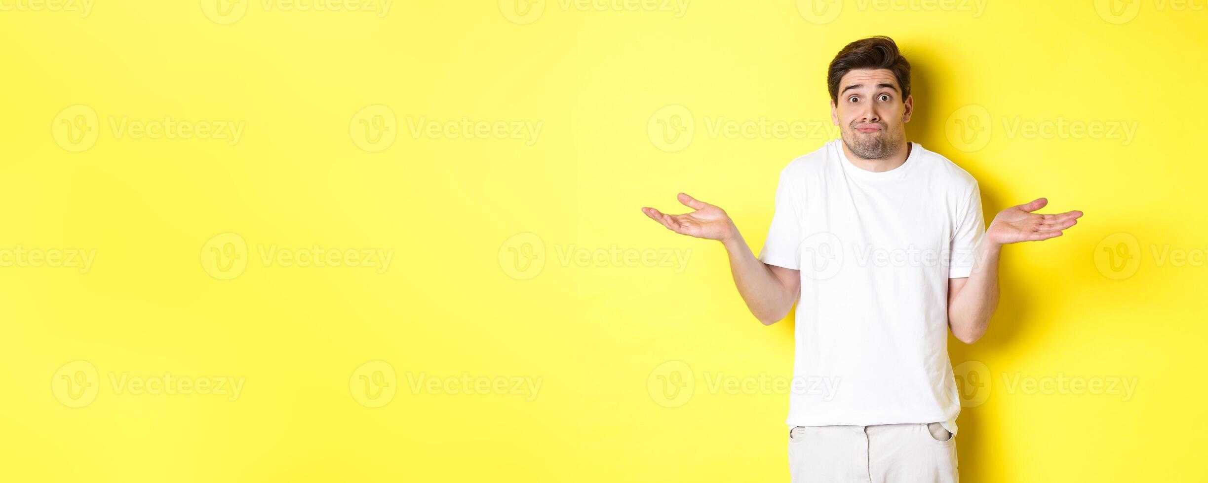Clueless man shrugging, spread hands sideways confused, dont know anything, standing over yellow background photo