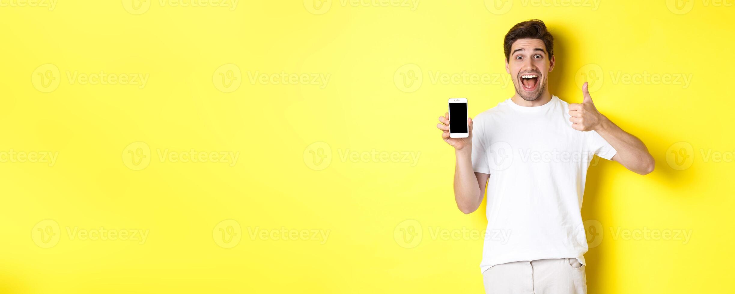 Happy young man showing thumb up and mobile phone screen, recommending application or internet website, standing over yellow background photo