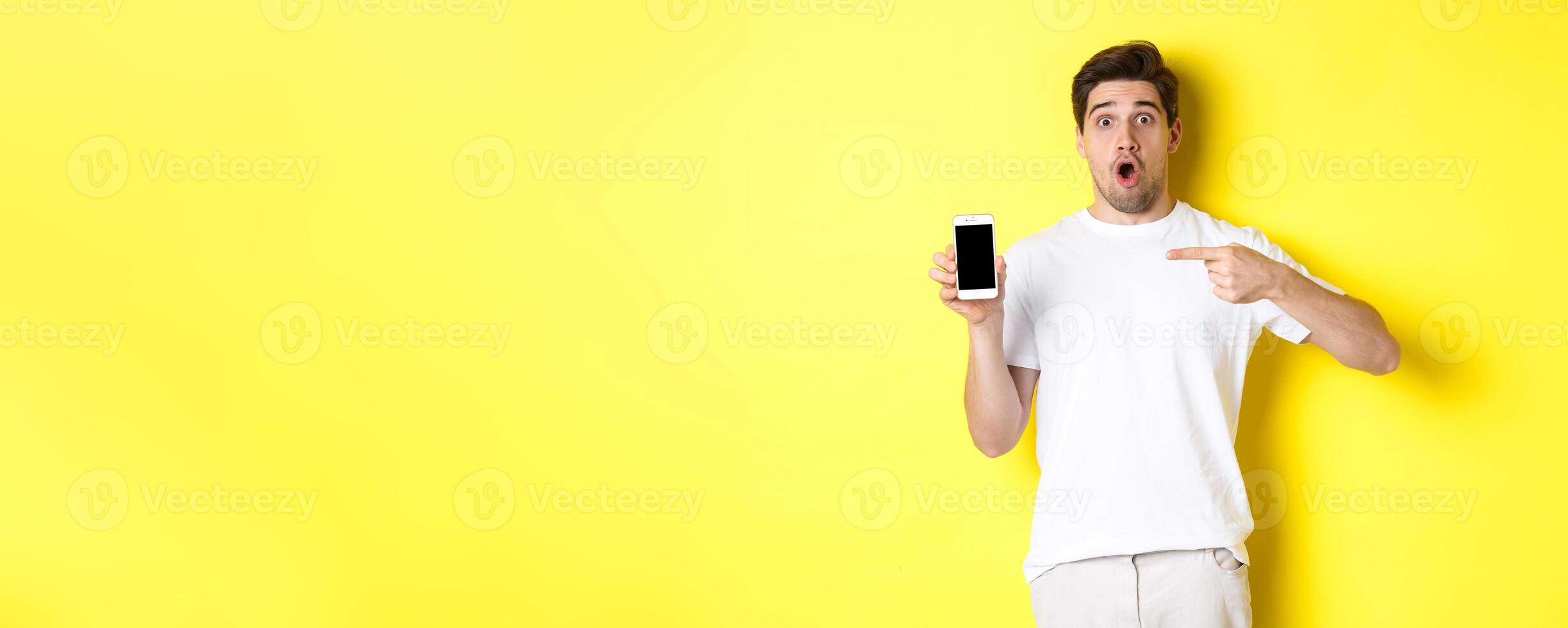 Impressed guy check out smartphone screen, pointing finger at display and gasping amazed, standing over yellow background photo