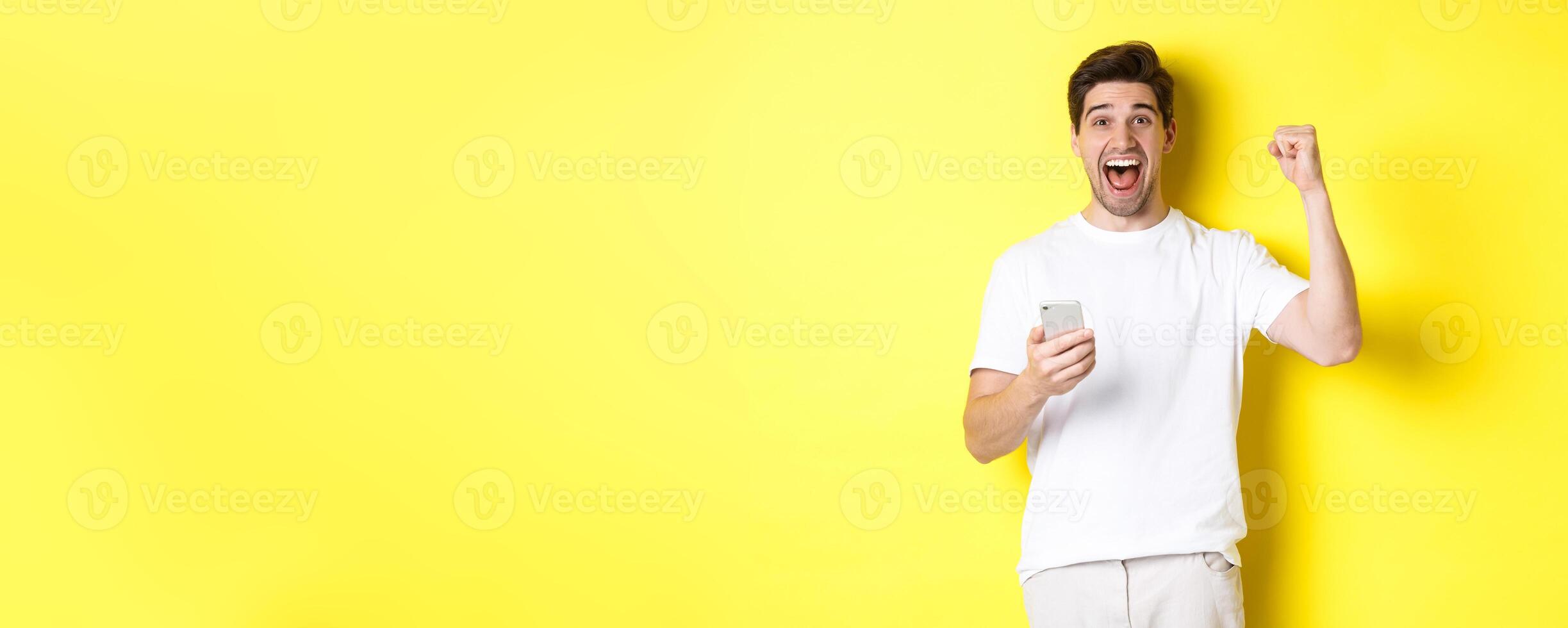 Cheerful man winning on smartphone, raising hand up and holding mobile, achieve app goal, standing over yellow background photo