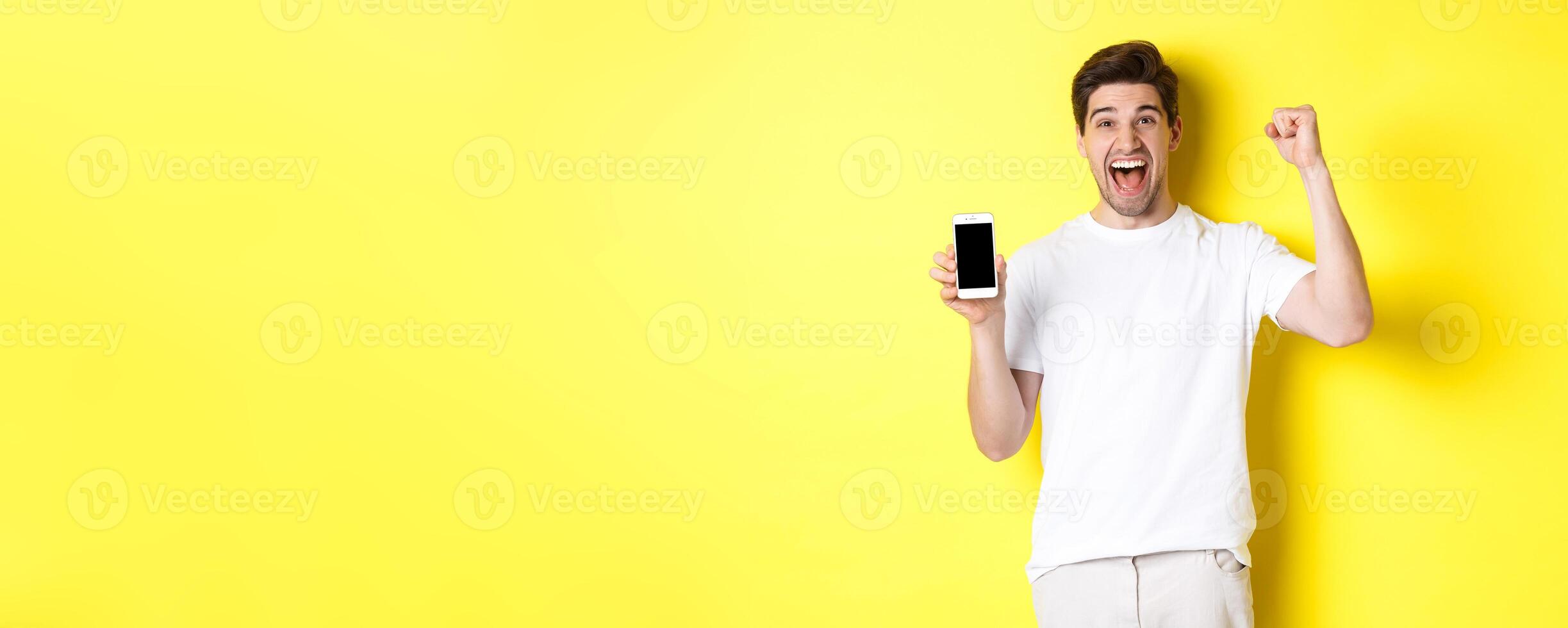 Cheerful guy showing smartphone screen, raising hand up and celebrating, triumphing over internet achievement, standing over yellow background photo