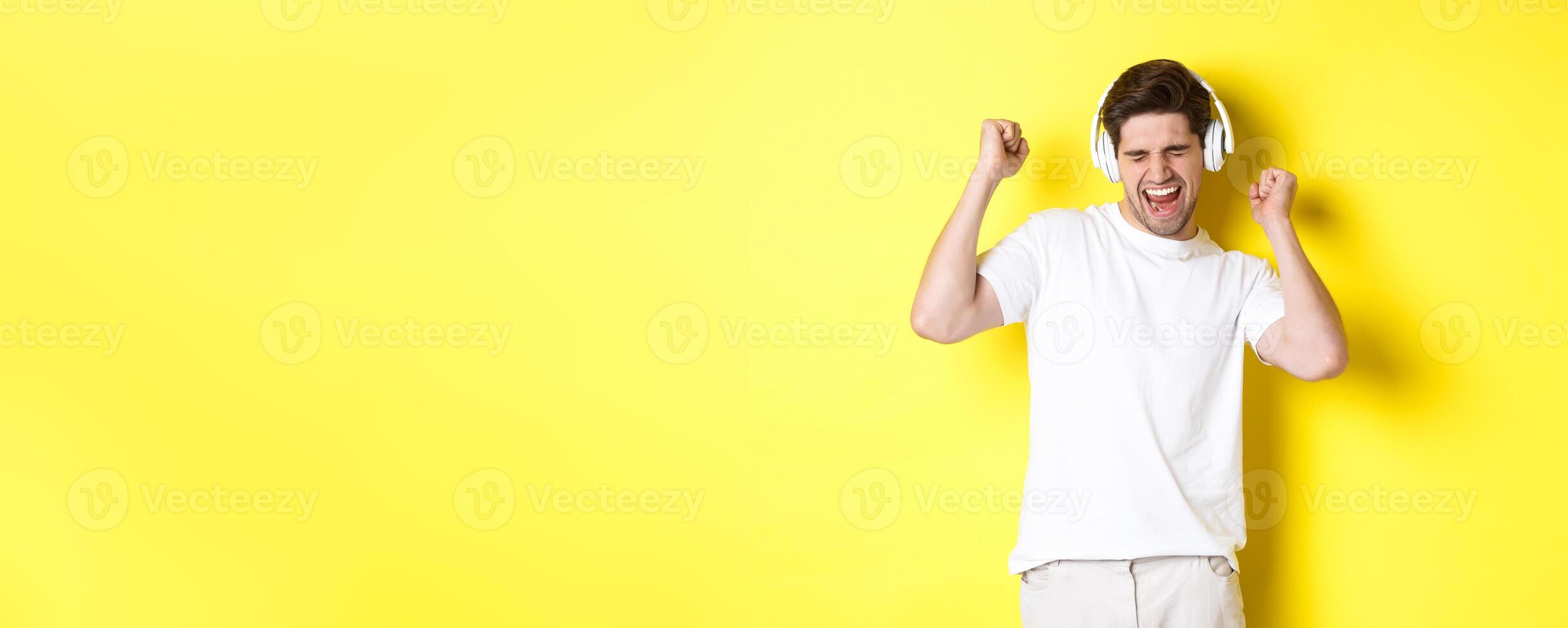 Happy man dancing and listening music in white headphones, standing over yellow background photo