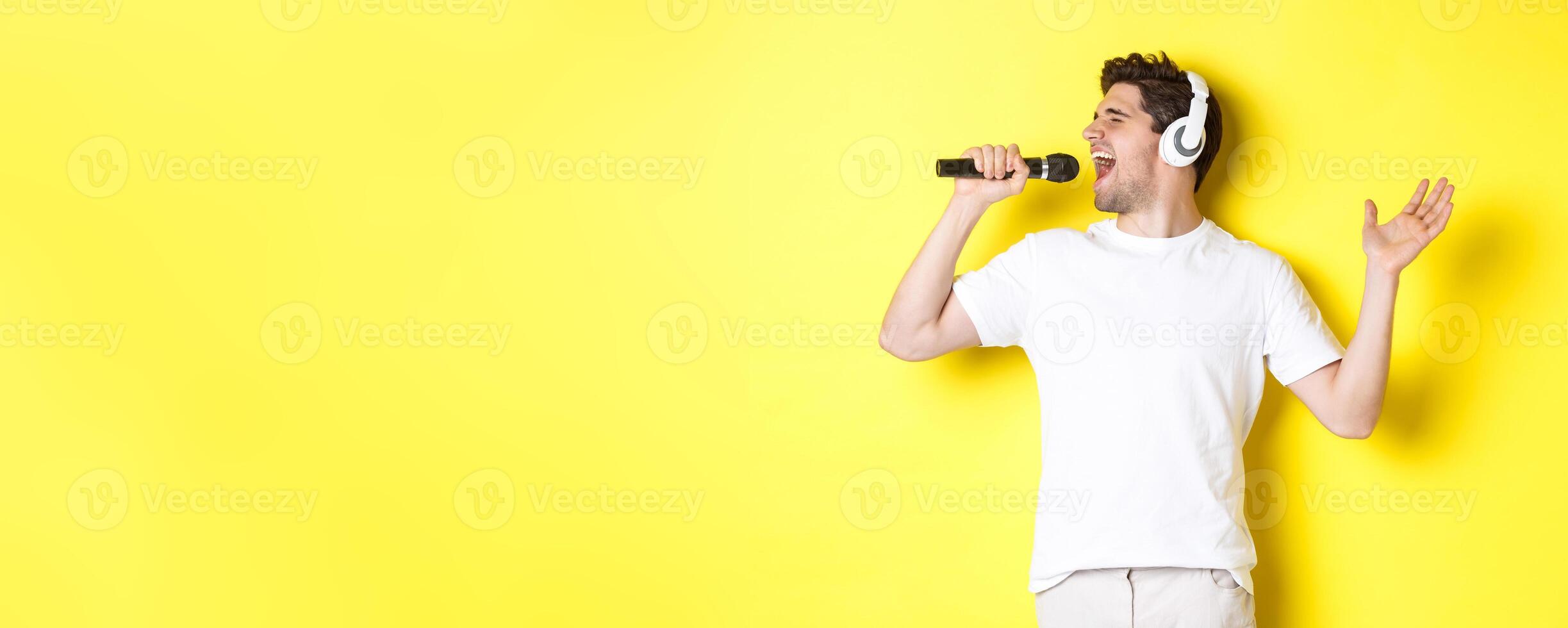 Passionate guy in headphones holding microphone, singing karaoke song, standing over yellow background in white clothes photo