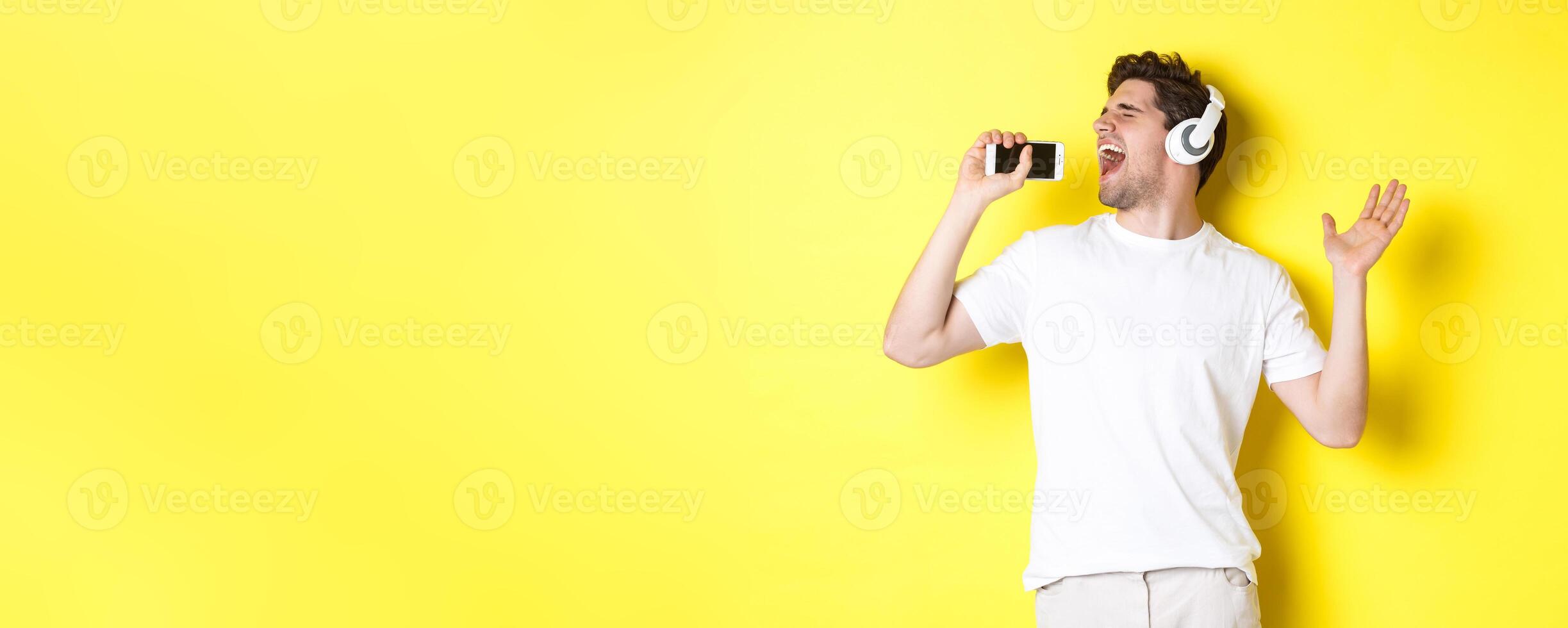 Happy guy playing karaoke app in headphones, singing into smartphone microphone, standing over yellow background photo
