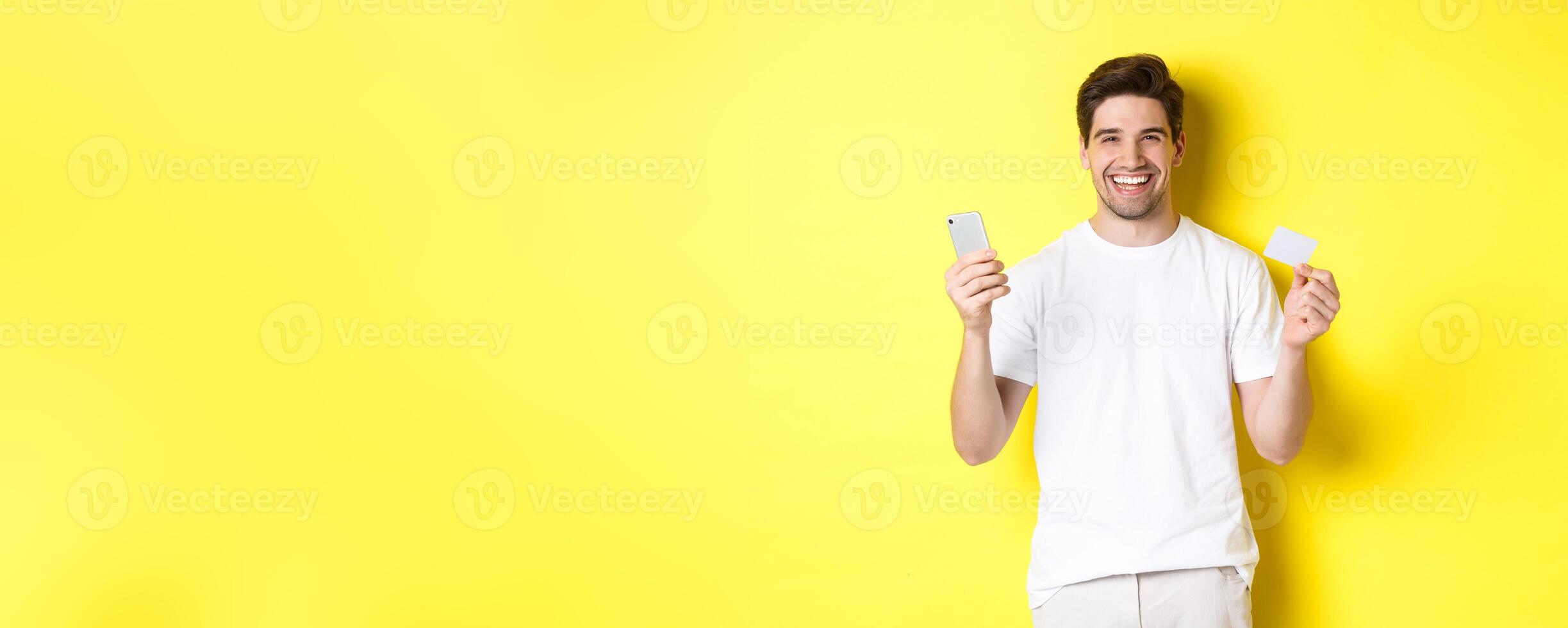 Happy young man shopping online in smartphone, holding credit card and smiling, standing over yellow background photo