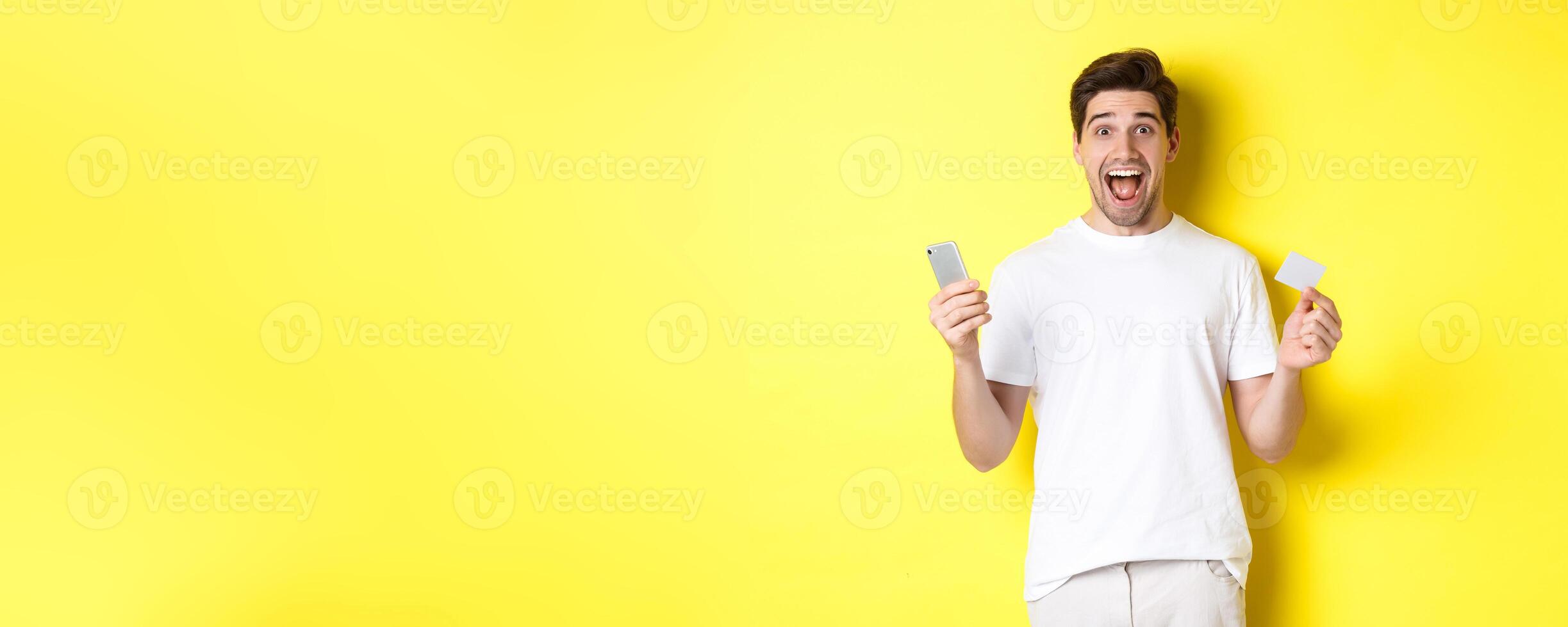 Surprised guy holding smartphone and credit card, online shopping on black friday, standing over yellow background photo