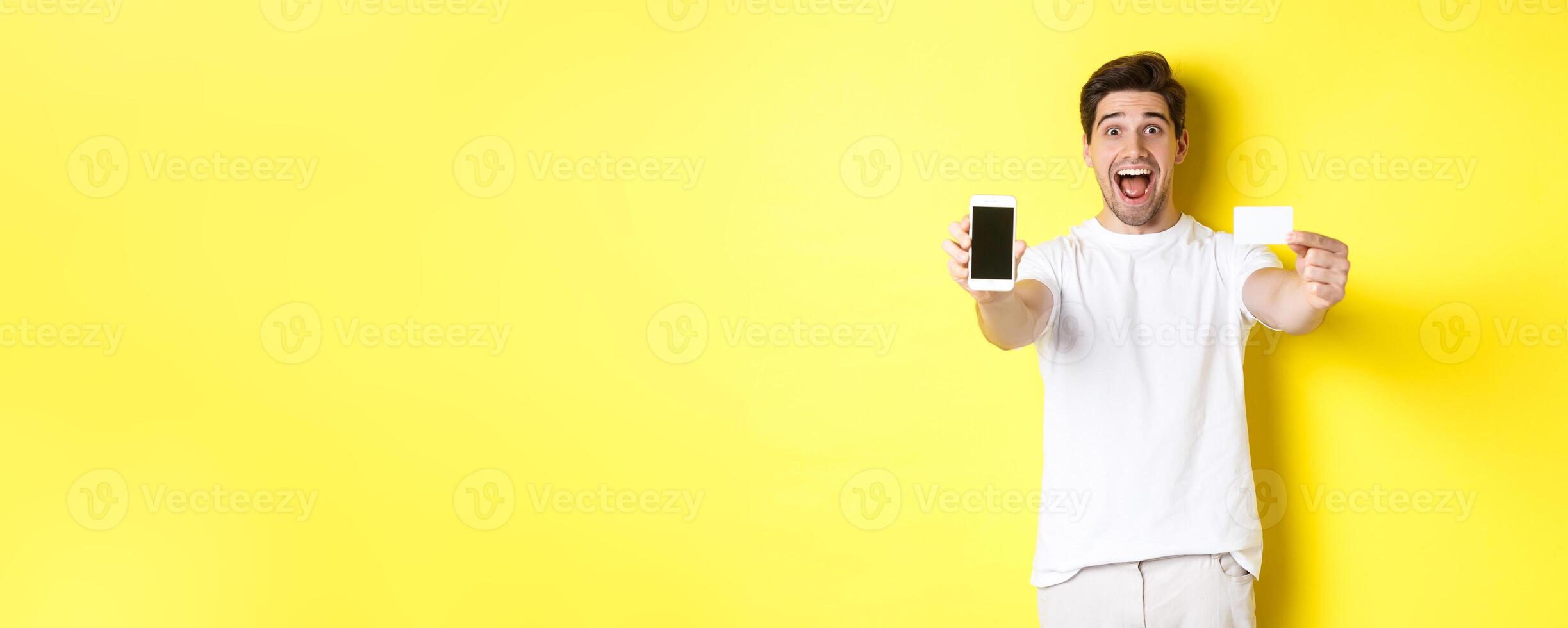 Handsome caucasian man showing smartphone screen and credit card, concept of mobile banking and online shopping, yellow background photo