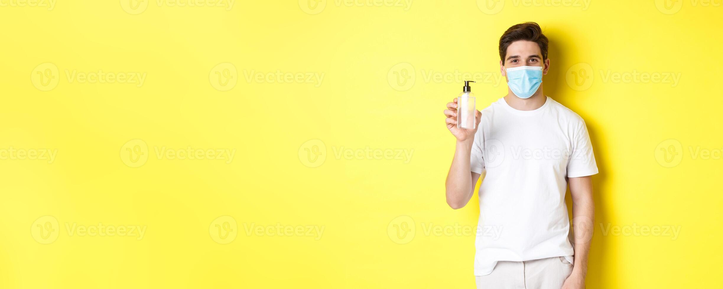 Concept of covid-19, quarantine and lifestyle. Young man in medical mask showing hand sanitizer, hands disinfection product, standing over yellow background photo