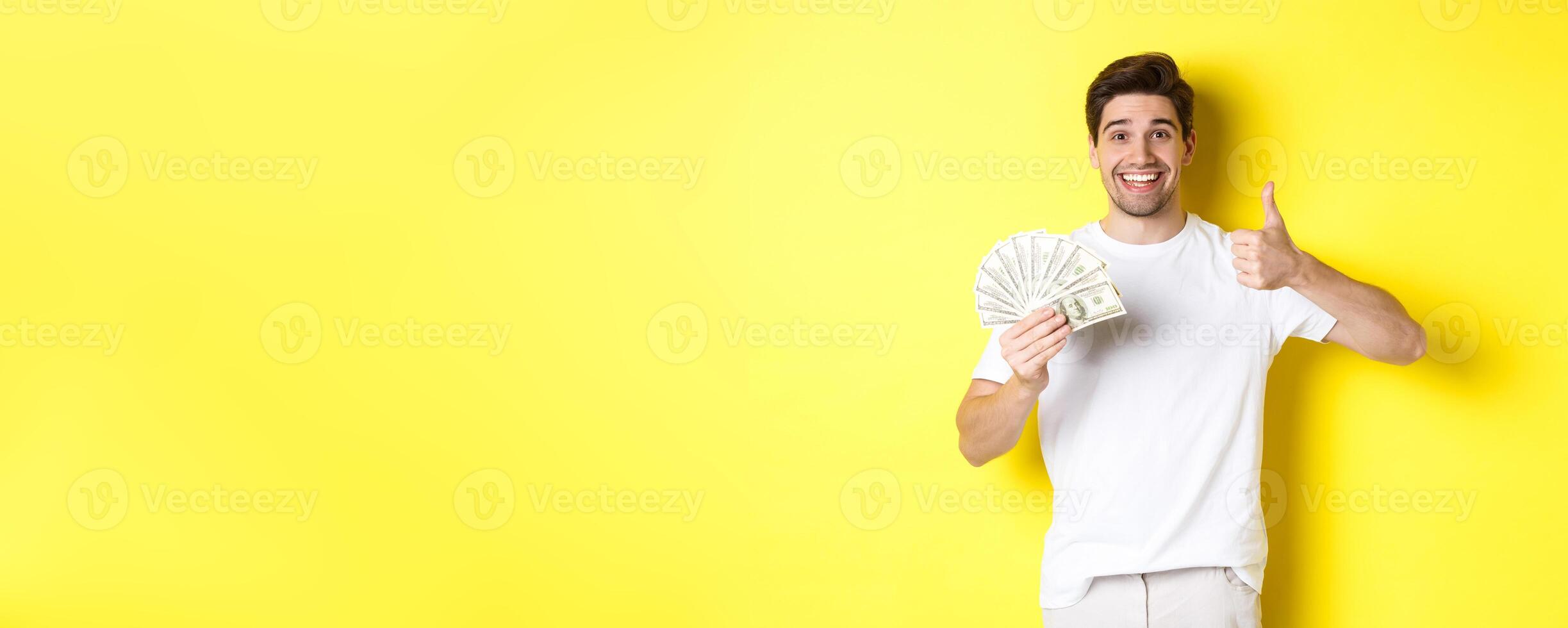 Satisfied buyer man showing thumb-up and holding money, shopping with cash, standing over yellow background photo