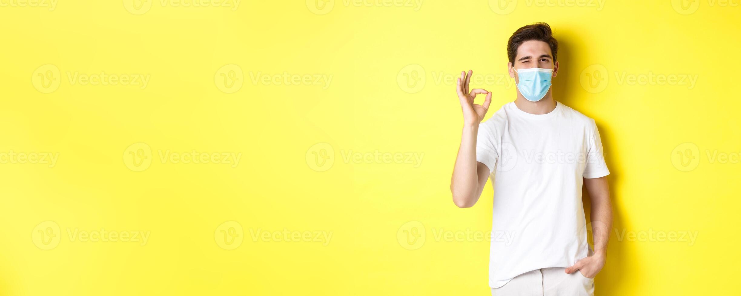 Concept of coronavirus, pandemic and social distancing. Confident young man in medical mask showing okay sign and winking, yellow background photo