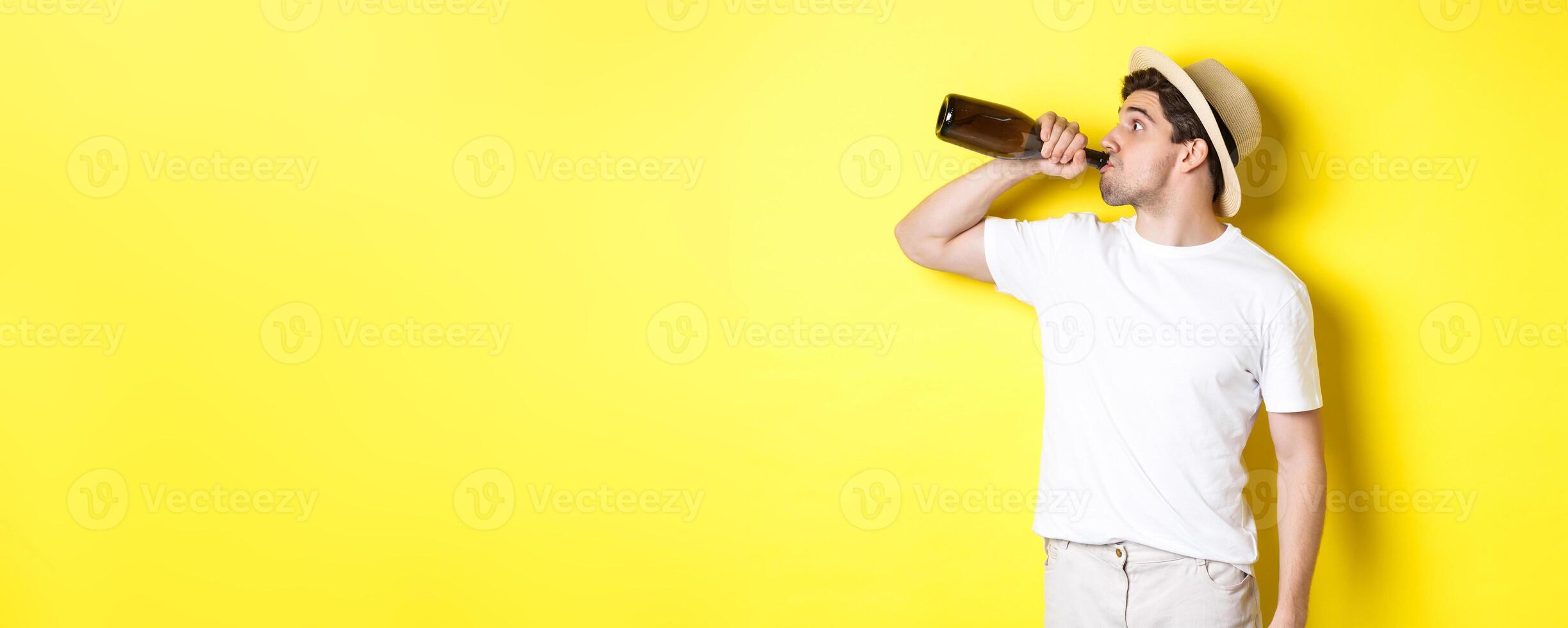 Concept of tourism and vacation. Man drinking wine from bottle on holidays, standing against yellow background photo