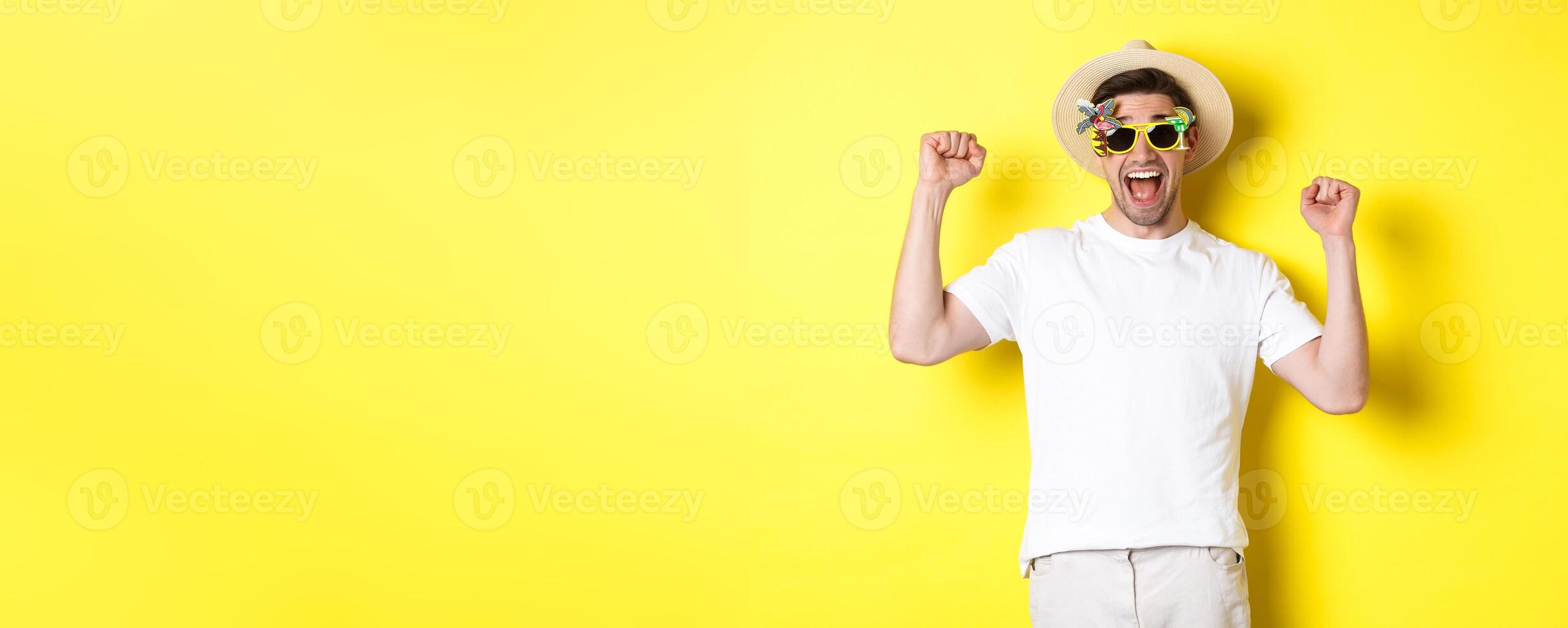Concept of tourism and lifestyle. Happy man winning trip to resort, shouting yes and raising hands up, triumphing, wearing sunglasses and summer hat, yellow background photo