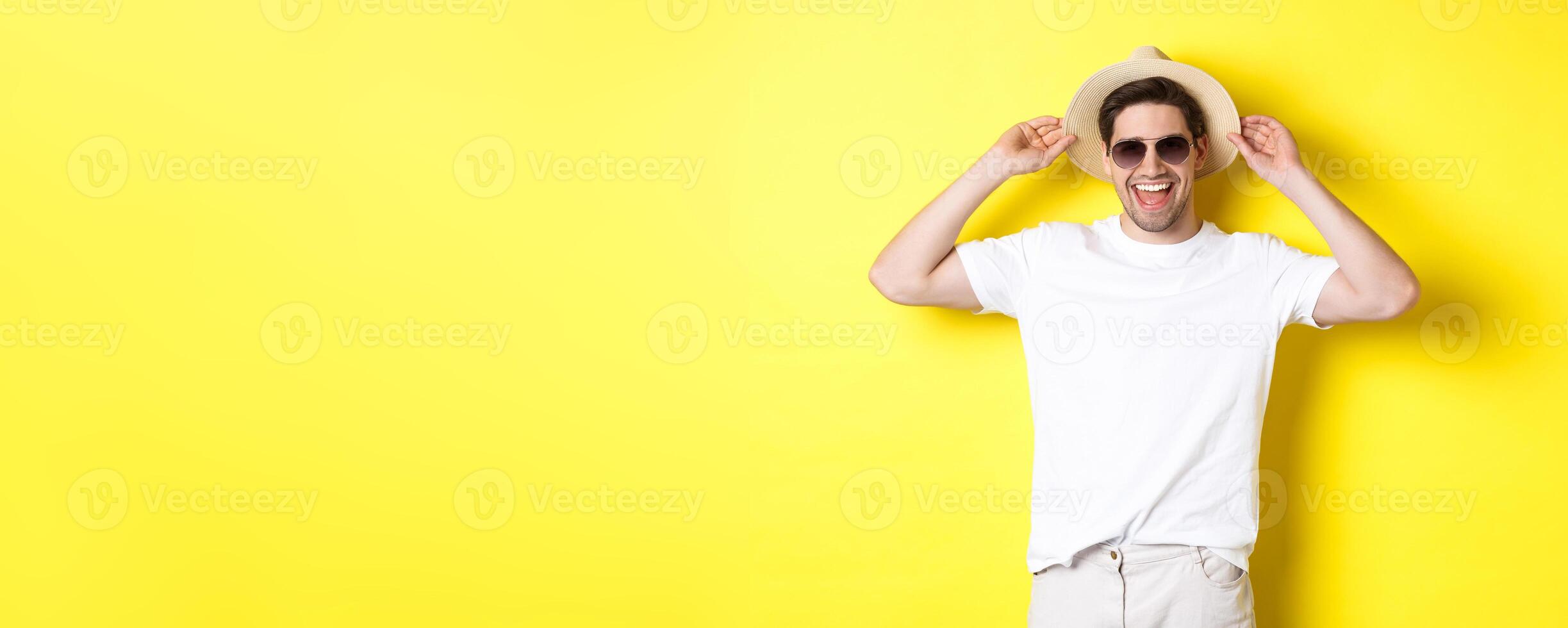 Happy man on vacation, wearing straw hat and sunglasses, smiling while standing against yellow background photo