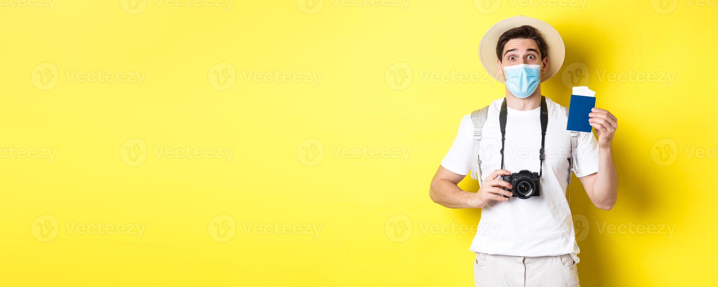 Concept of covid-19, travelling and quarantine. Happy man tourist with camera, showing passport and tickets for vacation, going on trip during pandemic, yellow background photo