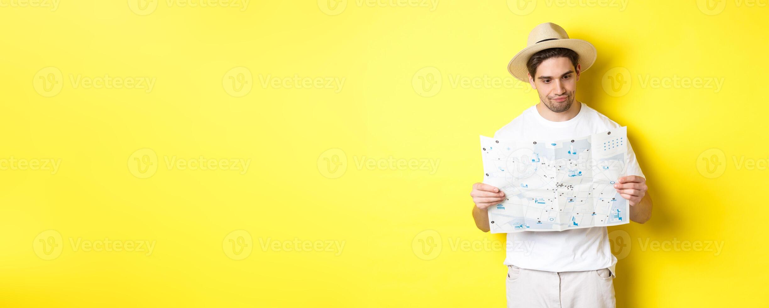 Travelling, vacation and tourism concept. Male tourist looking confused at map, looking for right direction, standing over yellow background photo