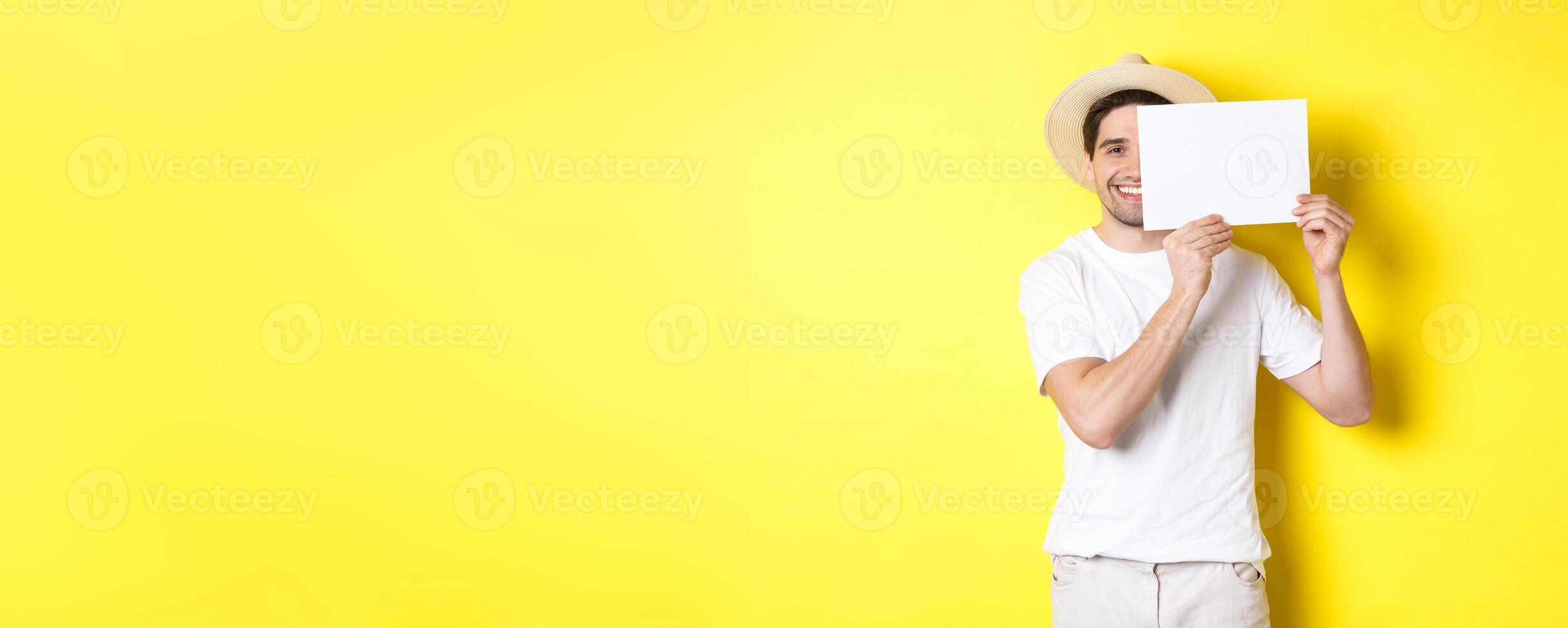Happy guy on vacation showing blank piece of paper for your logo, holding sign near face and smiling, standing against yellow background photo