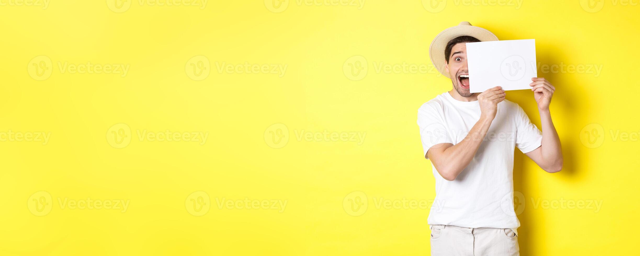 Excited tourist on vacation showing blank piece of paper for your logo, holding sign near face and smiling, standing against yellow background photo