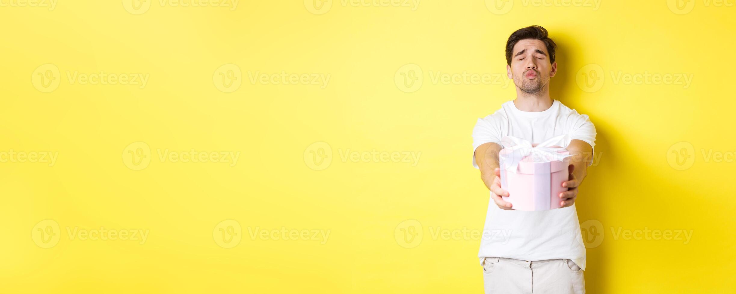 Concept of holidays and celebration. Boyfriend giving a gift, close eyes and pucker lips for kiss, standing over yellow background photo