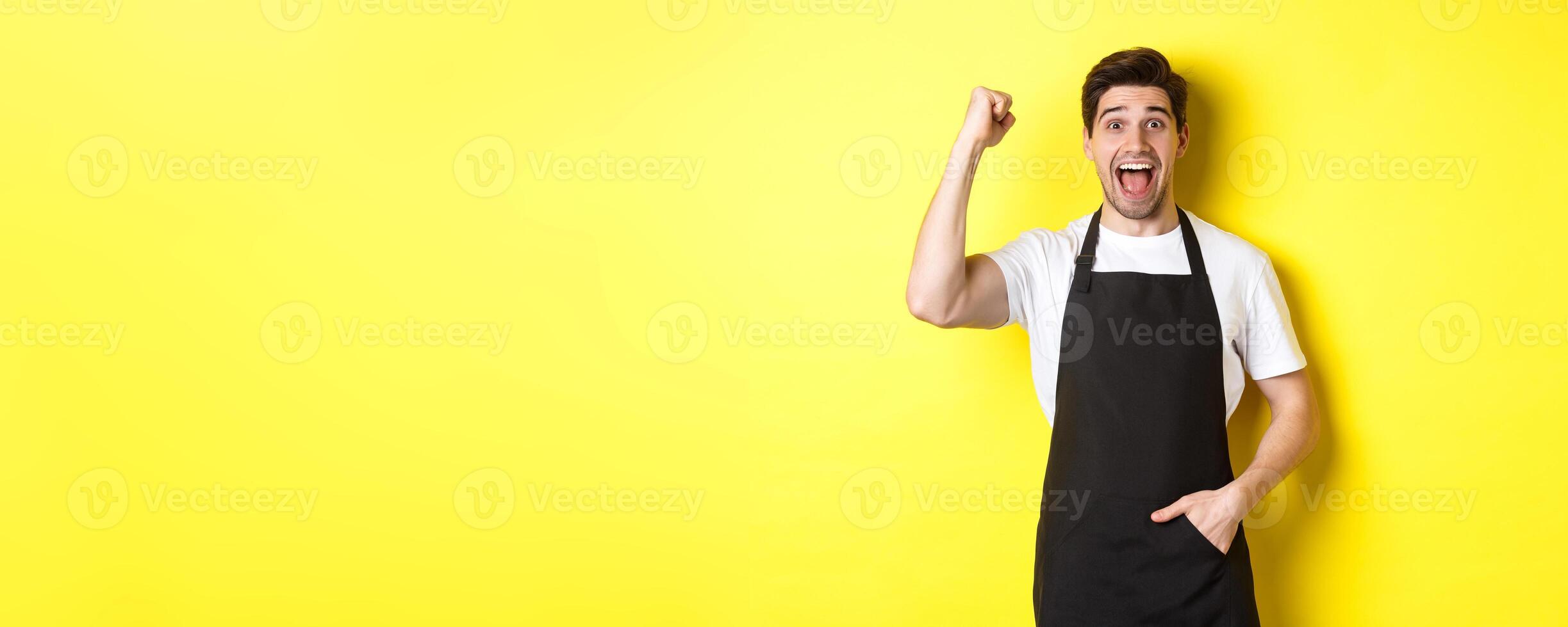 Cheerful seller making fist pump, rejoicing and triumphing, standing in black apron against yellow background photo