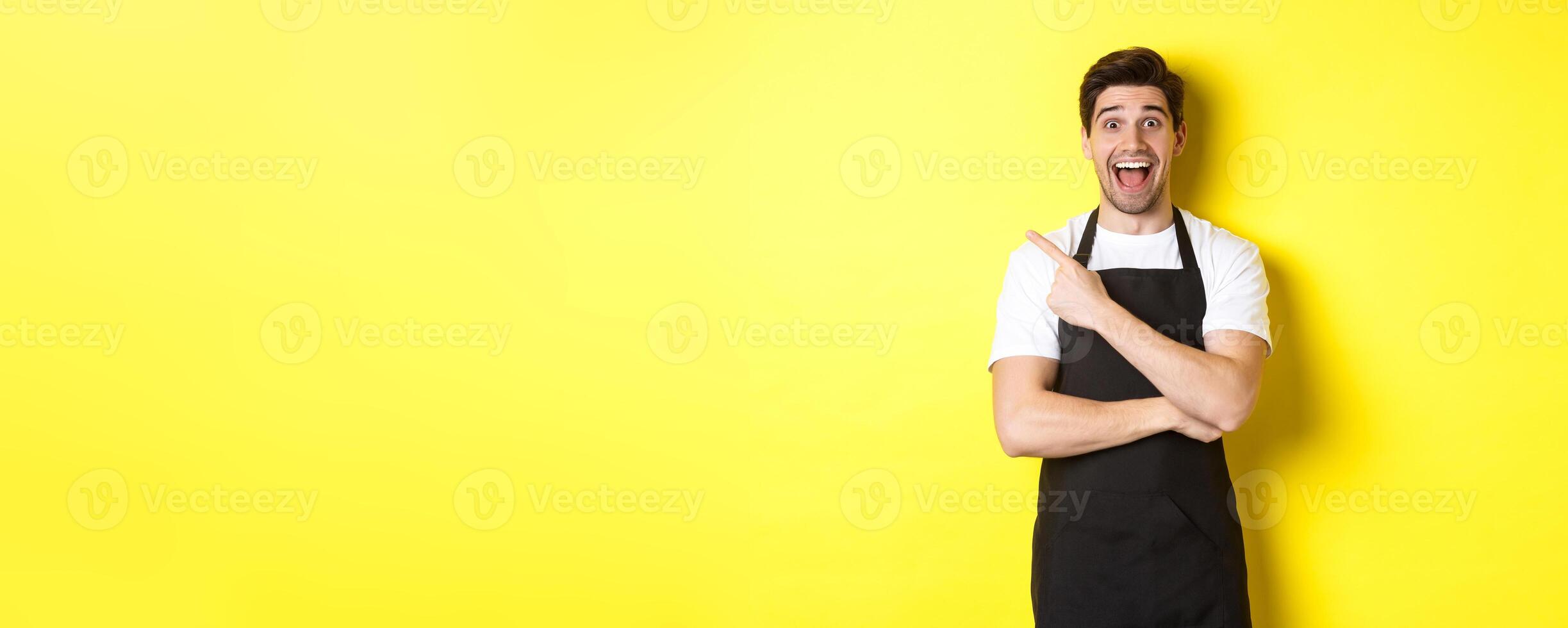Surprised salesman in black apron pointing finger upper left corner, showing shop promo offer, standing against yellow background photo