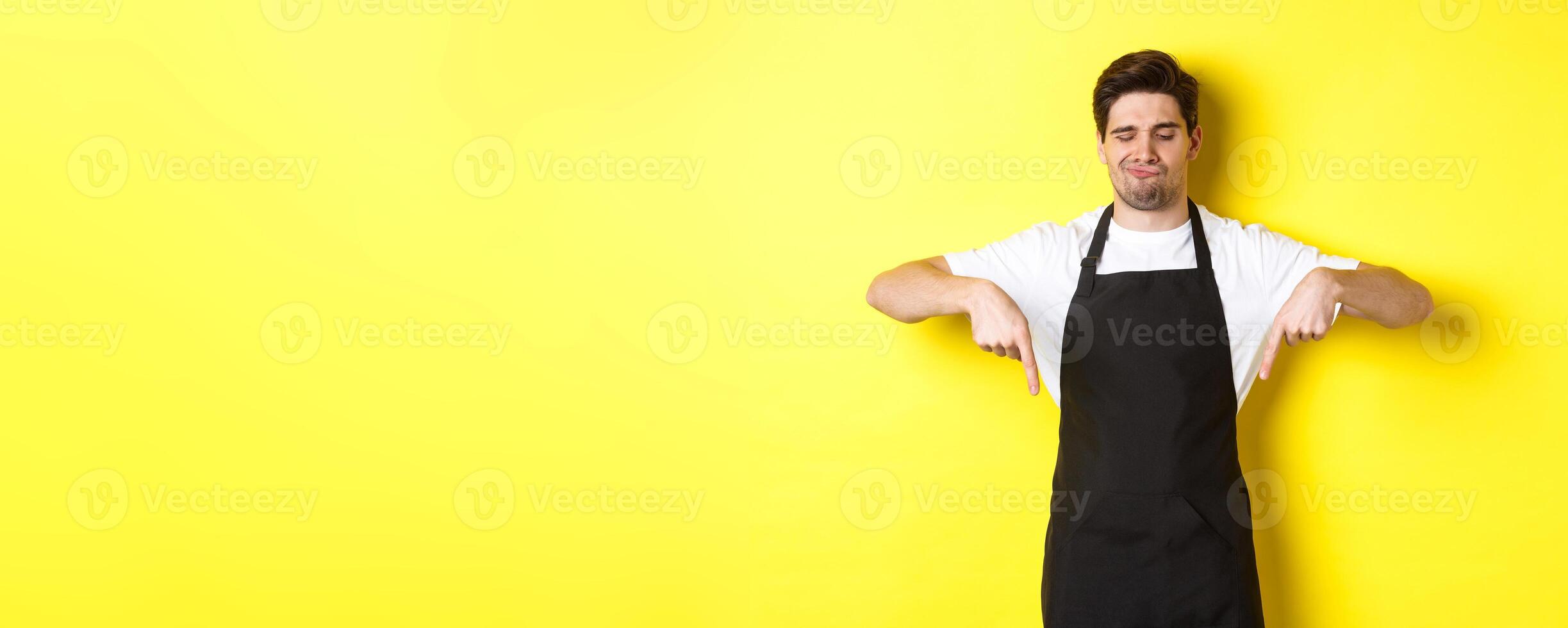 Doubtful barista complaining, pointing fingers down and grimacing displeased, standing in black apron against yellow background photo