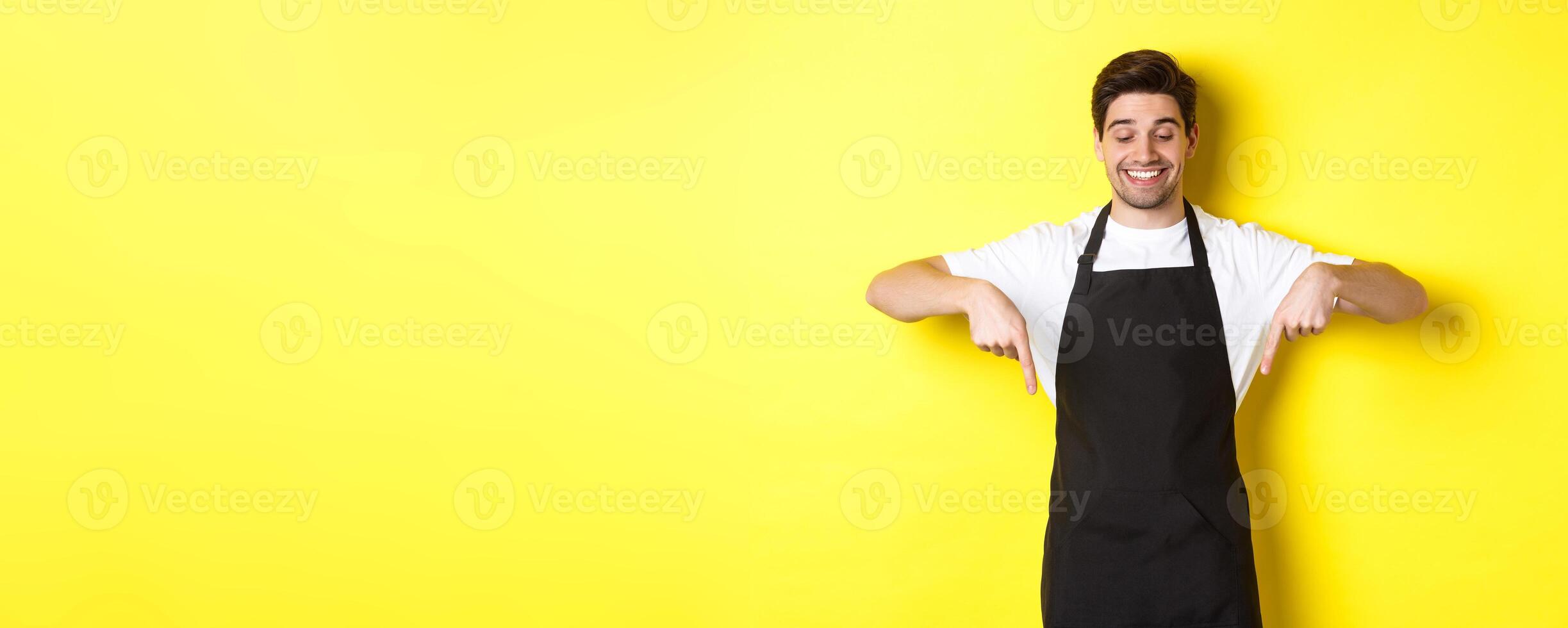 Happy coffee shop worker in black apron pointing fingers down, looking pleased and smiling, showing logo, standing over yellow background photo