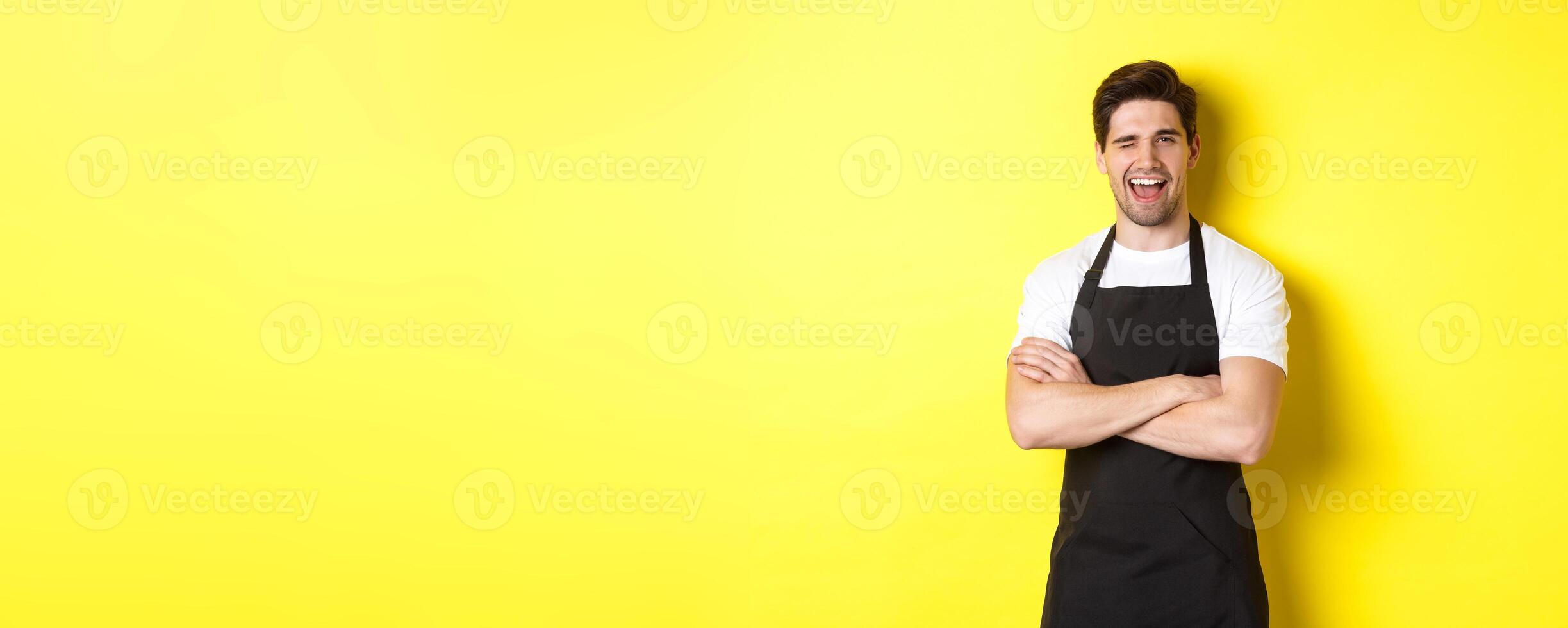Handsome barista in black apron winking at you, wearing black apron uniform, standing over yellow background photo