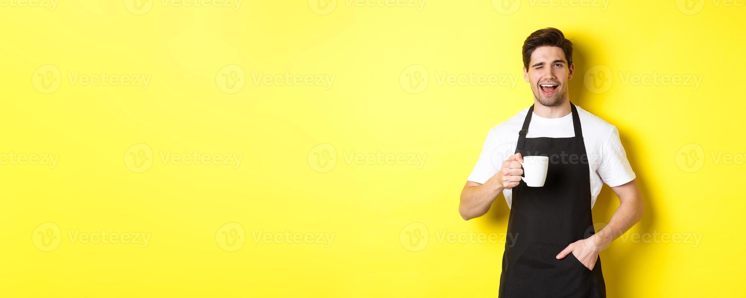 Cheerful barista in black apron serving coffee and winking, standing against yellow background photo