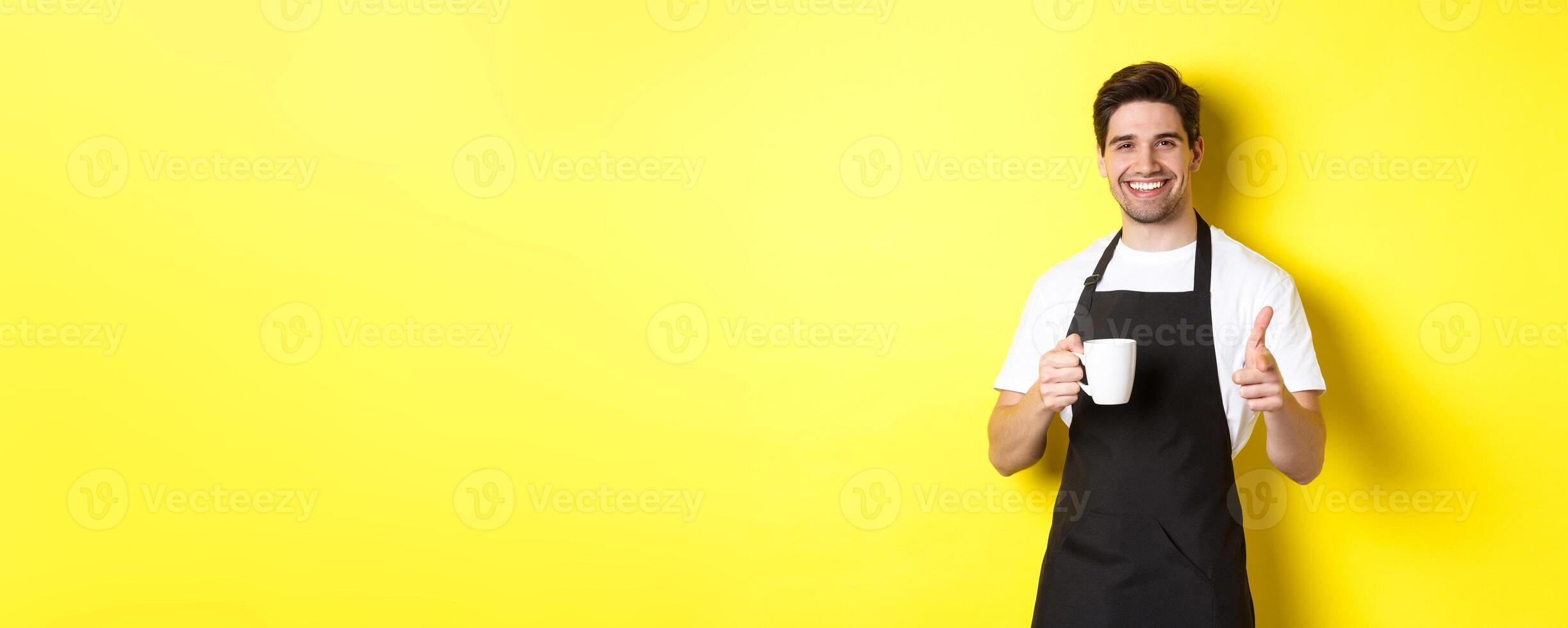 Barista bringing coffee and pointing finger gun at camera, standing in black apron against yellow background photo