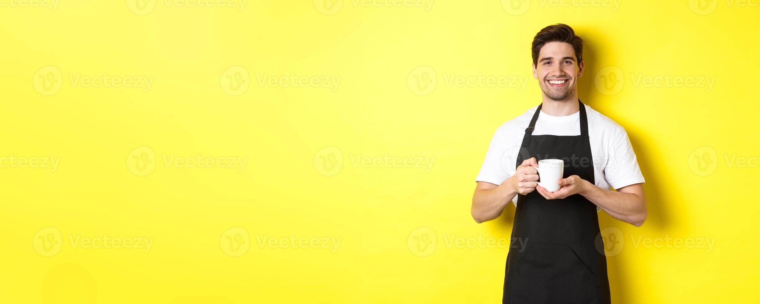 Handsome barista serving coffee, bring cup, standing in black apron with friendly smile photo
