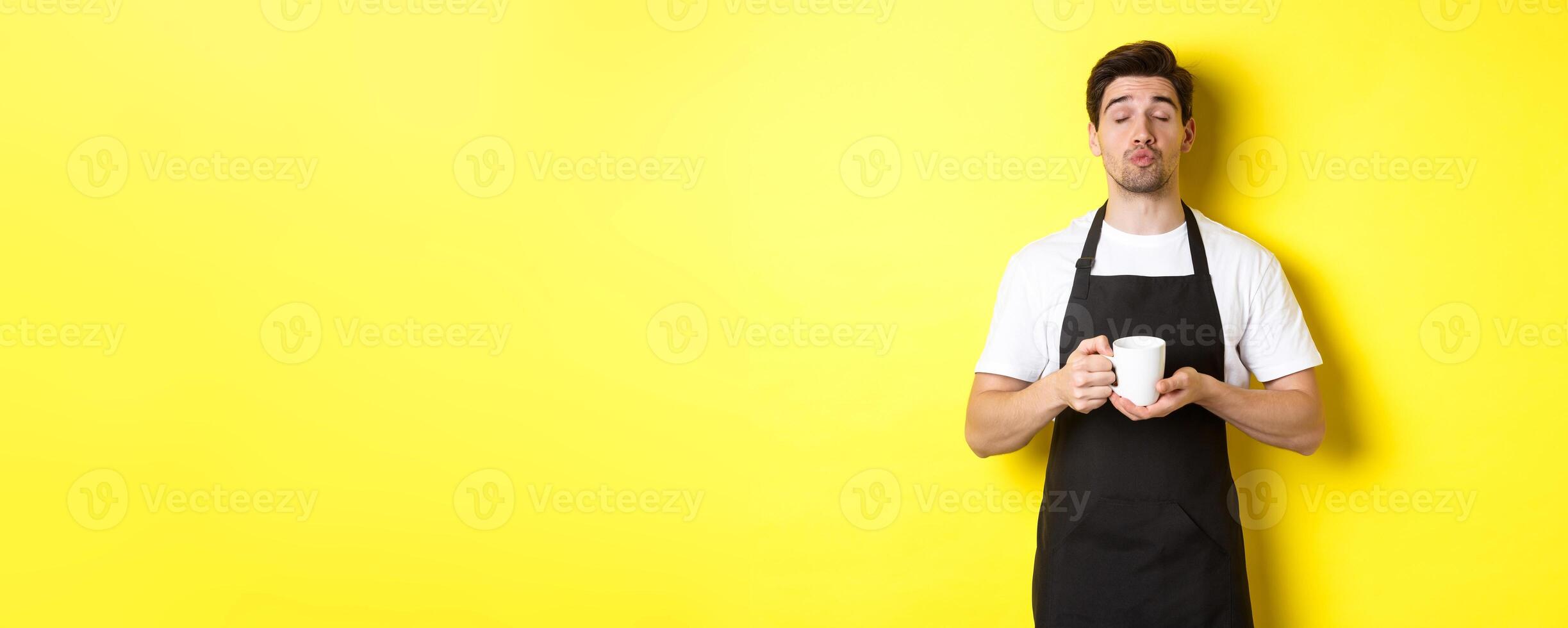 Man in black apron bringing cup of coffee and waiting for kiss, standing over yellow background photo