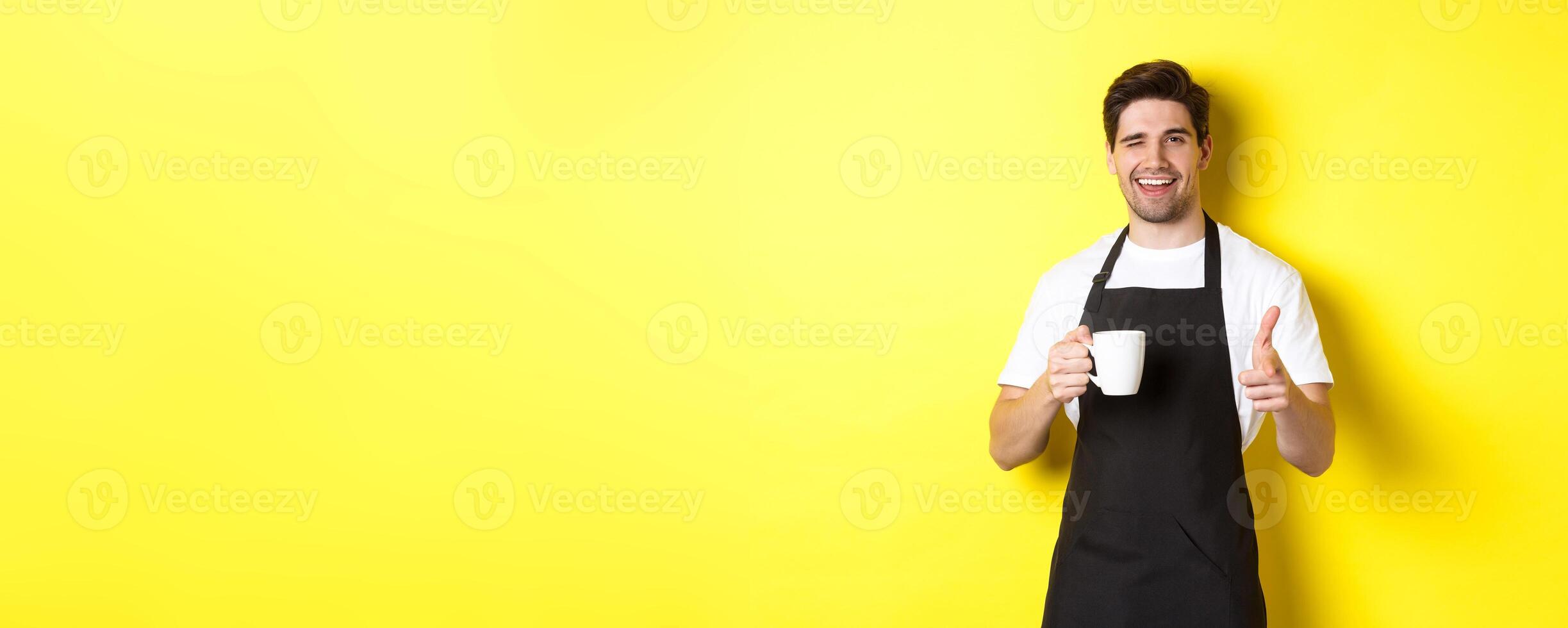 Handsome barista in black apron holding coffee cup, pointing finger at you, inviting visit cafe, standing over yellow background photo