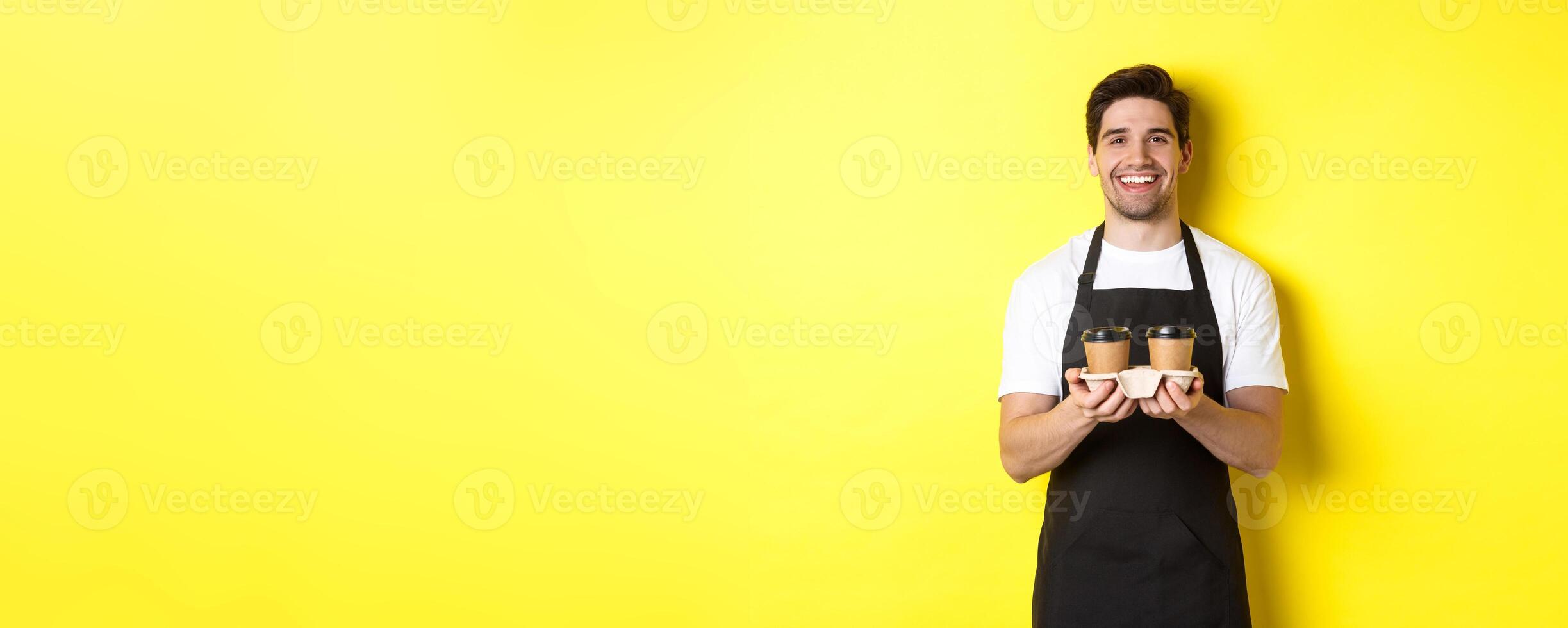 Friendly barista in black apron giving takeaway order, holding two cups of coffee and smiling, standing over yellow background photo